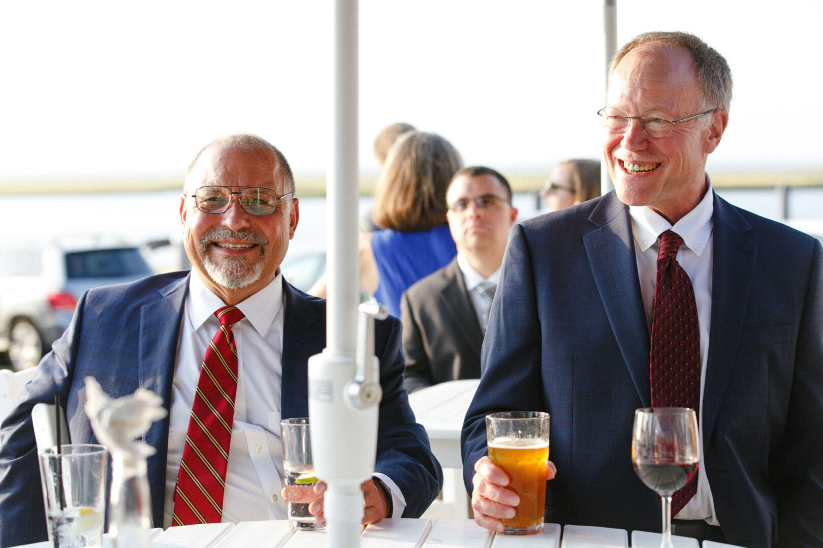 Two men in suits, one sporting a red tie, holding drinks and smiling at an outdoor gathering, exuding the joy and camaraderie typical of a Stone Harbor Yacht Club wedding celebration.