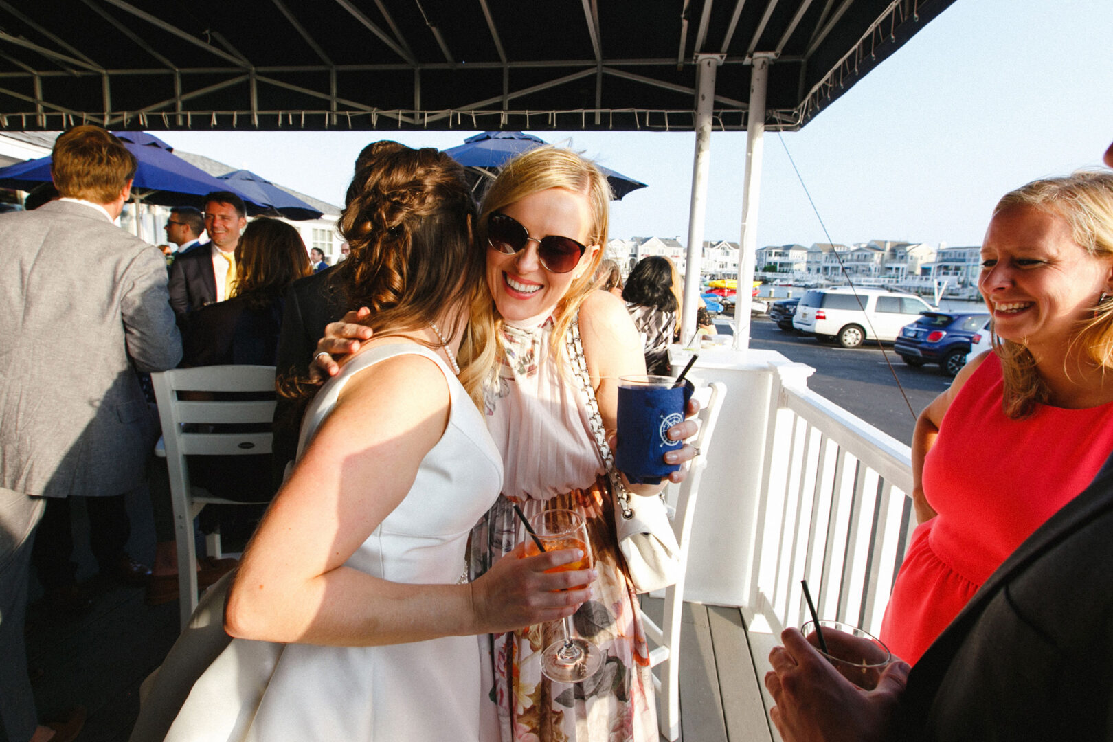 Woman in a white dress hugs a smiling woman with sunglasses holding a drink on the deck of Stone Harbor Yacht Club during a lively wedding celebration, others mingling happily in the background.