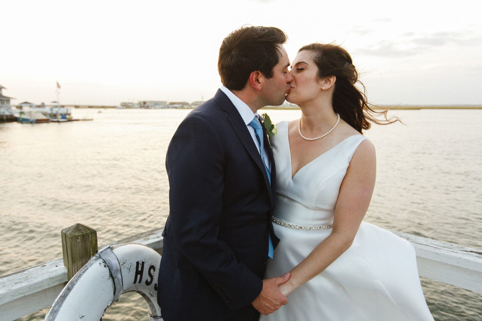Dressed in elegant formal attire, a couple shares a kiss on the dock beside a life preserver, with the tranquil water and sky as their backdrop—a perfect moment from their Stone Harbor Yacht Club wedding.