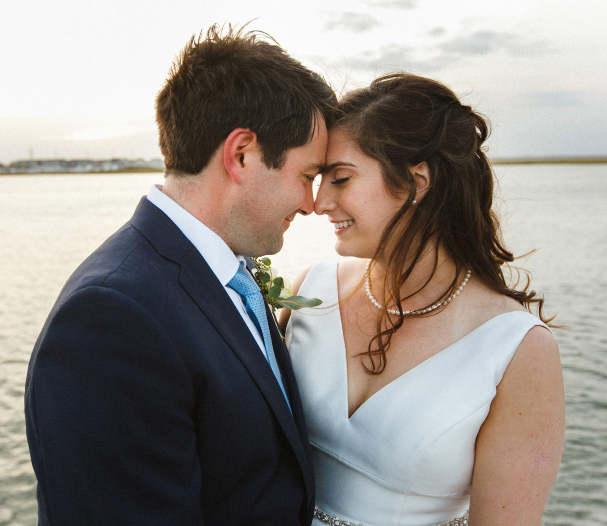 A couple in formal attire embraces by the water at Stone Harbor Yacht Club, touching foreheads and smiling softly. The background features a calm body of water and a light sky, capturing the essence of their intimate wedding day.