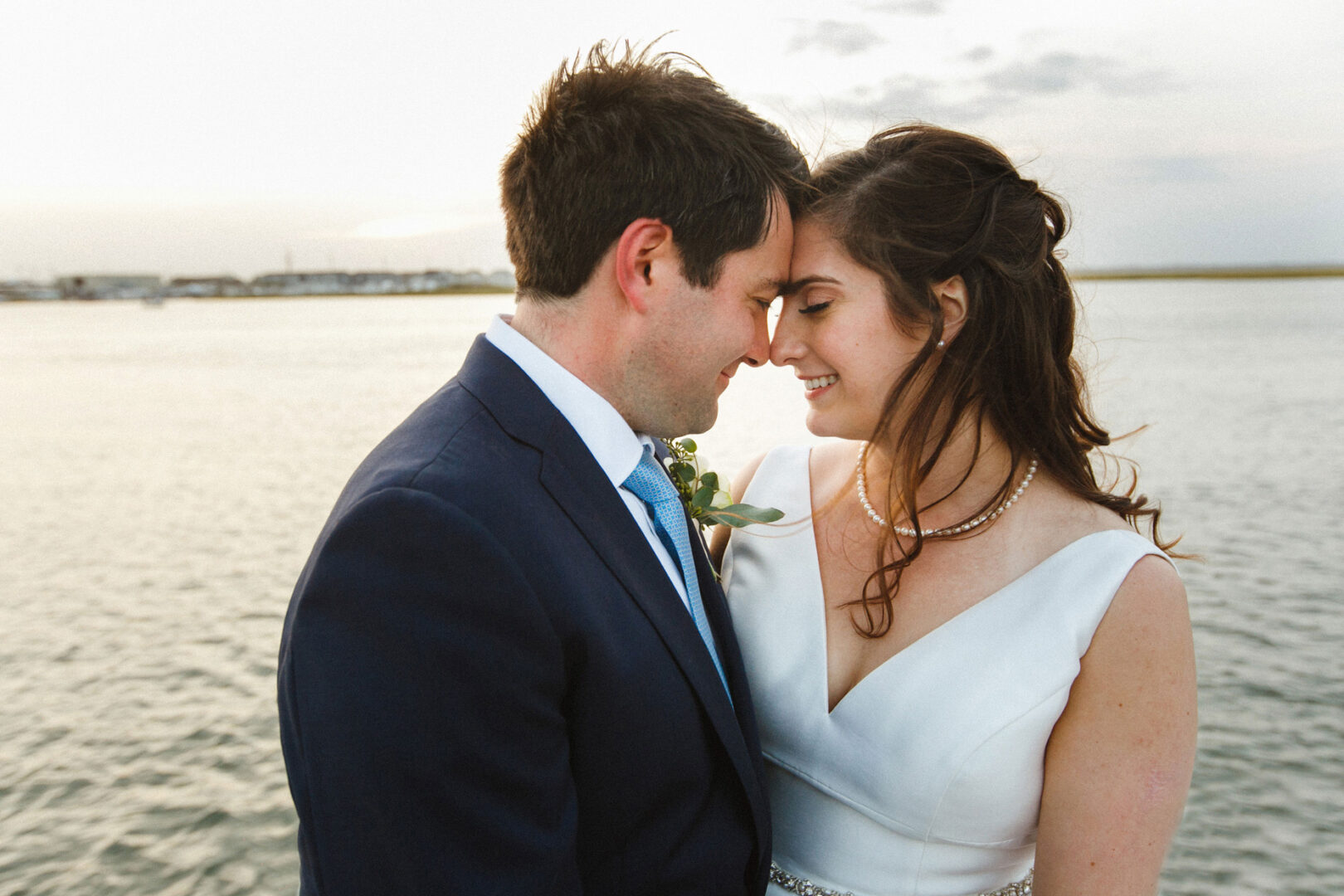 A couple in formal attire embraces by the water at Stone Harbor Yacht Club, touching foreheads and smiling softly. The background features a calm body of water and a light sky, capturing the essence of their intimate wedding day.