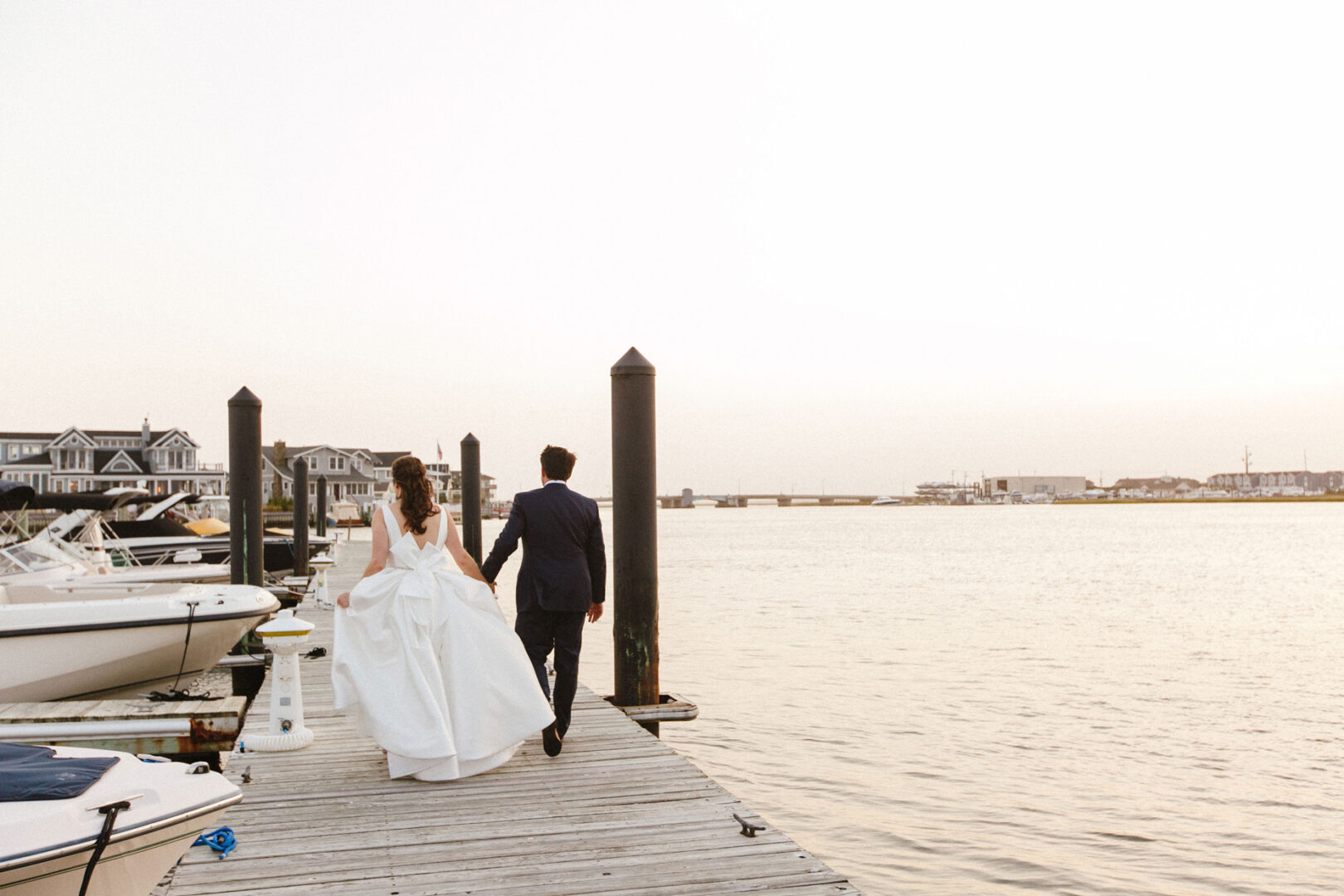 The bride and groom walk hand in hand on a wooden pier beside boats, with houses and a calm body of water at their Stone Harbor Yacht Club wedding.