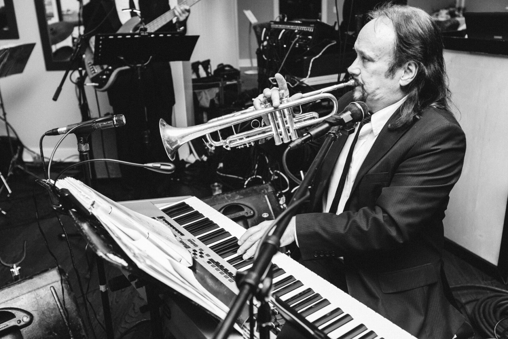 During a live performance at the Stone Harbor Yacht Club Wedding, a musician in a suit masterfully plays the trumpet and keyboard simultaneously, captivating the audience with his unique talent.