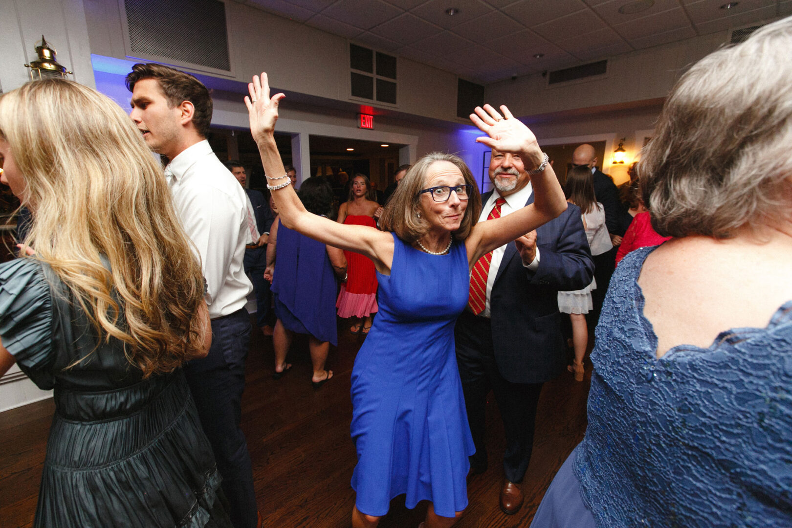 At a Stone Harbor Yacht Club wedding, a woman in a blue dress twirls gracefully, her arms raised amidst the lively crowd in the elegant indoor event space.