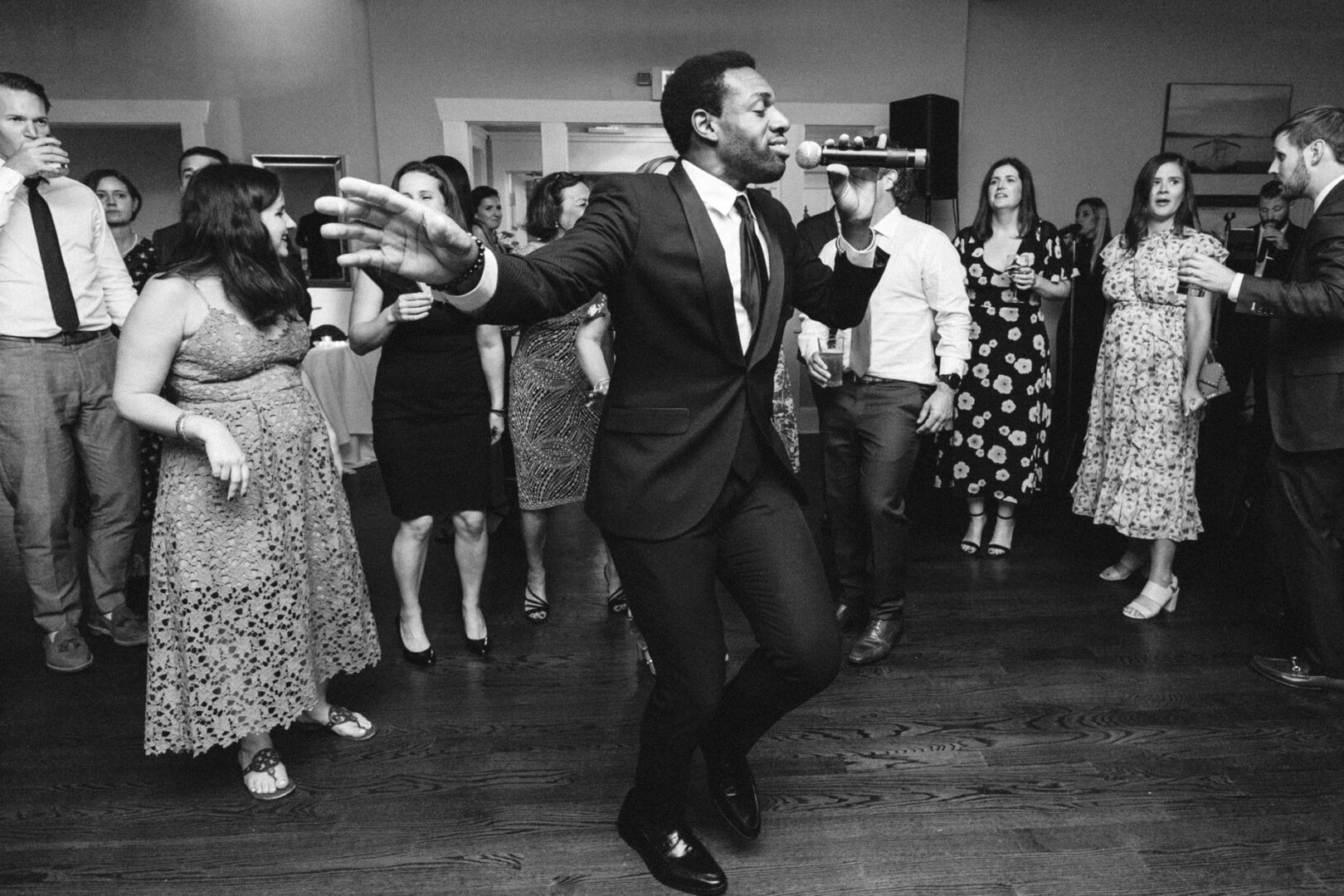 At a Stone Harbor Yacht Club wedding, a man in a suit sings into a microphone while dancing. A group of people stands around him, clapping and enjoying the performance at the elegant event.
