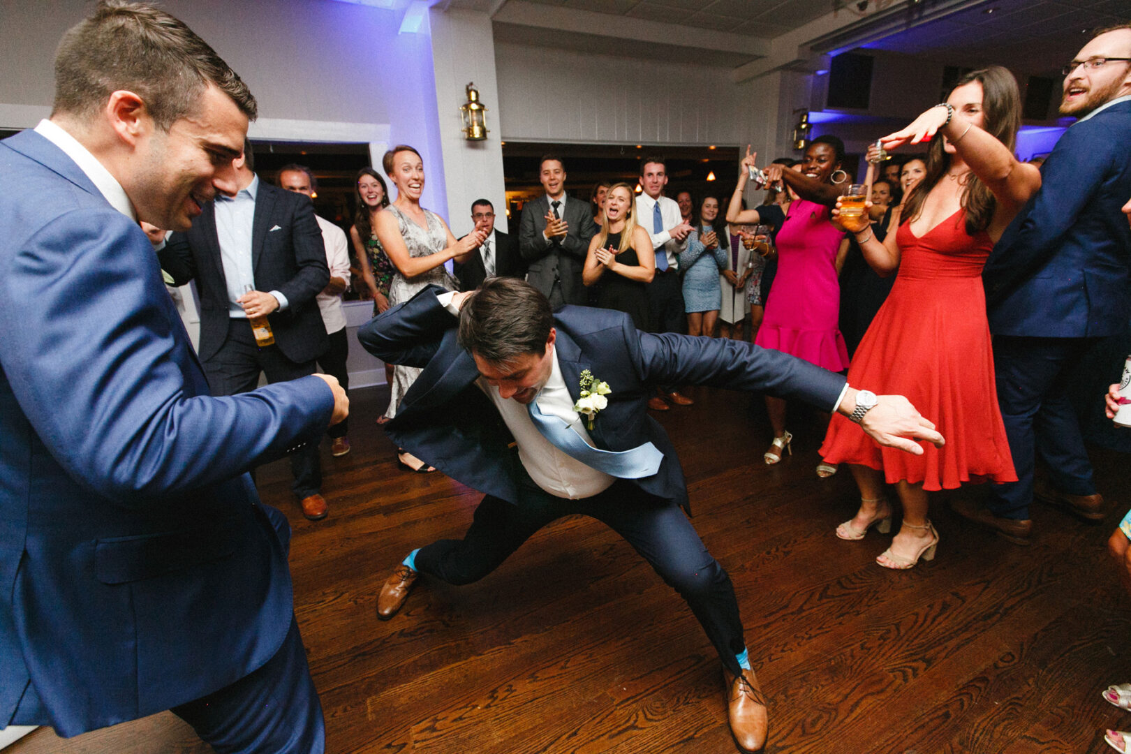 At a lively Stone Harbor Yacht Club wedding, a man in a suit performs an energetic dance move on the wooden floor, surrounded by a cheering crowd in festive attire.