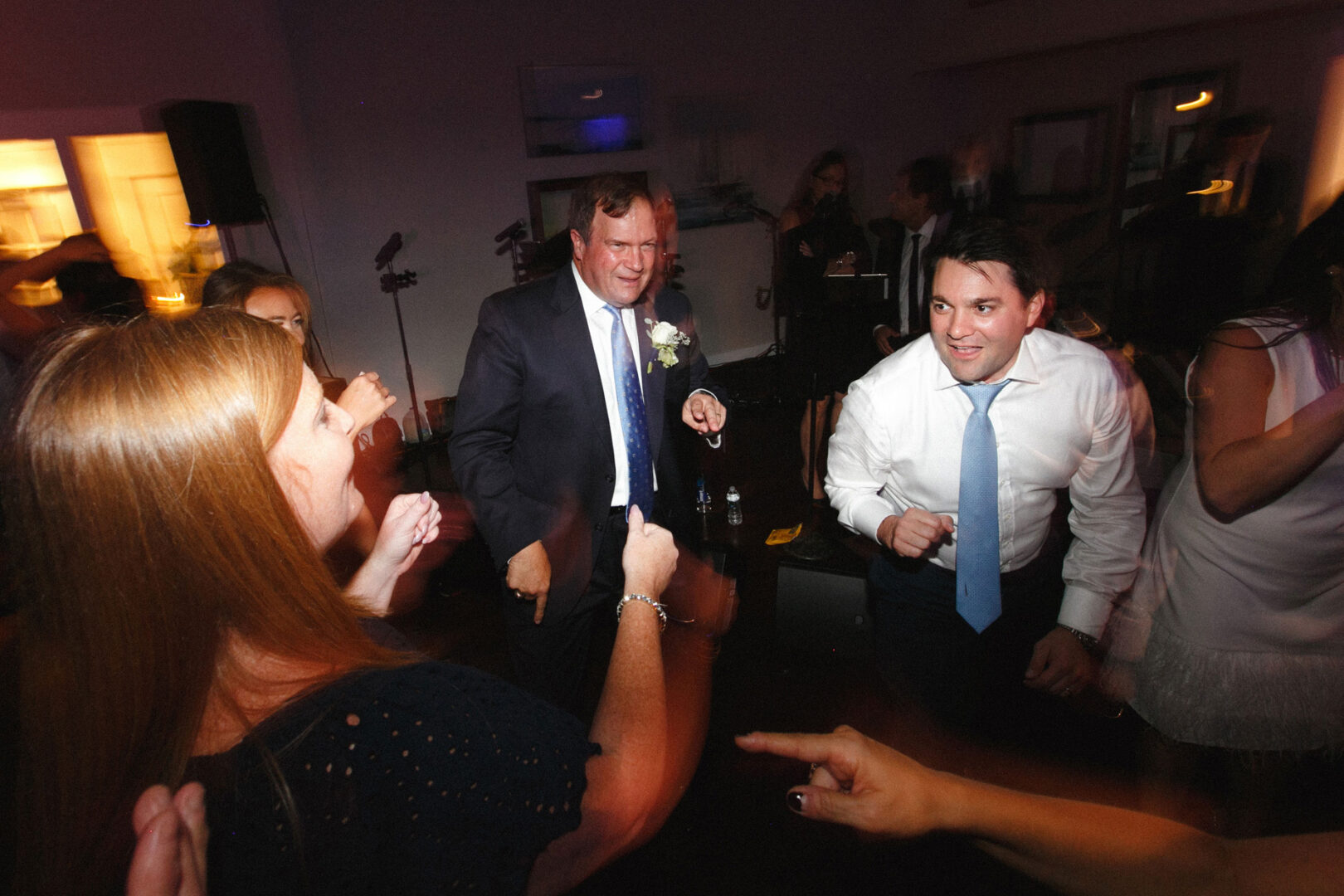 Guests in formal attire are dancing gracefully at an indoor event, reminiscent of a Stone Harbor Yacht Club wedding. The dimly lit room casts a romantic ambiance, with background figures softly blurred in the elegant setting.