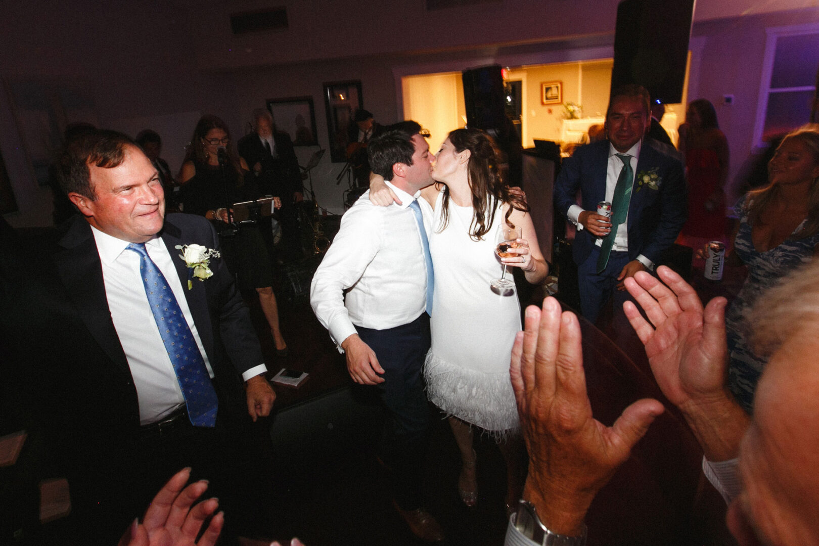 A bride and groom kiss on the dance floor of the Stone Harbor Yacht Club, surrounded by clapping guests in a warmly lit room.