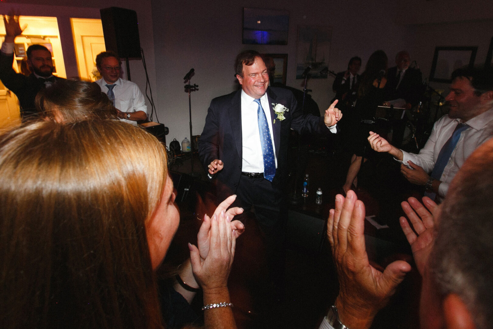 A man in a suit dances and smiles among the crowd at the Stone Harbor Yacht Club wedding. People around him are clapping and taking photos, capturing the joyful celebration.