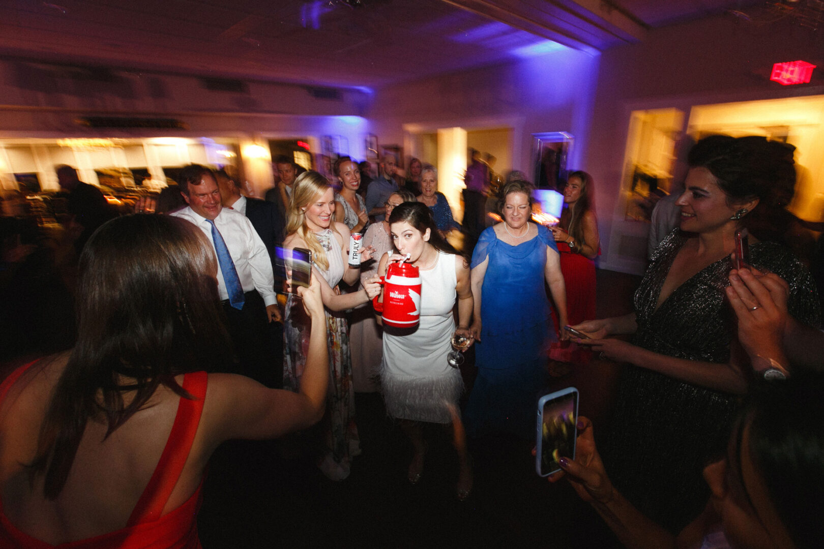 In the dimly lit room of the Stone Harbor Yacht Club wedding, colorful lights spark excitement. One person holds a red cup while others capture the joyous moments on their phones. The festive mood is palpable as everyone dances with delight.