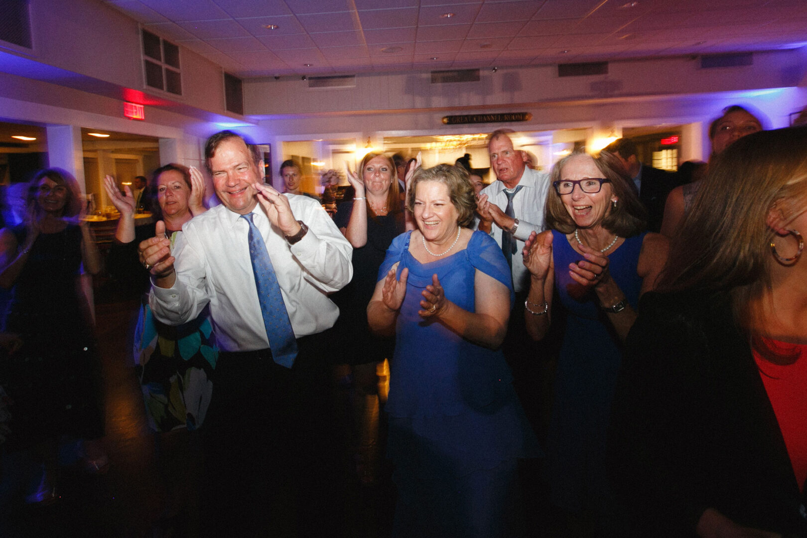 In a brightly lit room reminiscent of a joyous Stone Harbor Yacht Club wedding, a group, including a man and woman in the foreground, are clapping and smiling.