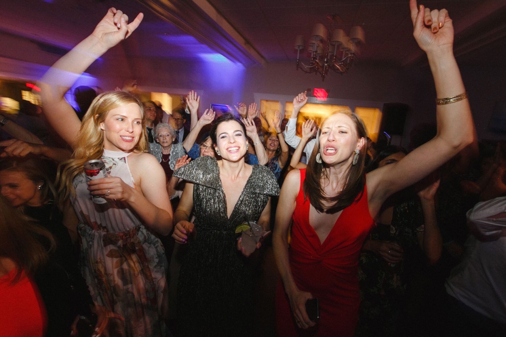 In the dimly lit room of Stone Harbor Yacht Club's wedding celebration, a group of people danced energetically. In the foreground, two women raised their hands and smiled, lost in the joyful rhythm of the evening.