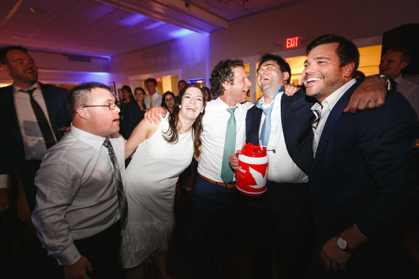 A group of people in formal attire, perhaps from a Stone Harbor Yacht Club wedding, laugh and hold each other while standing indoors. One person joyfully holds a red container, adding a splash of color to the celebration.