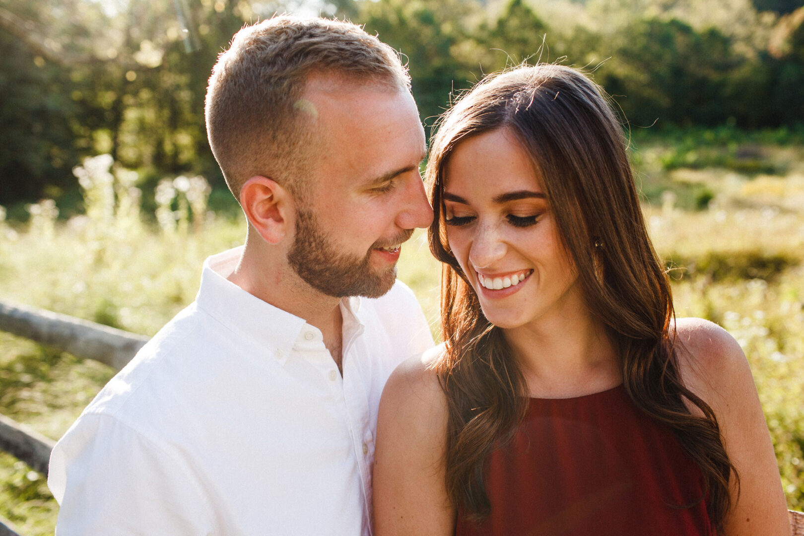 In a sunlit garden reminiscent of a Valley Forge Engagement Session, a couple stands closely together, smiling warmly. The man wears a crisp white shirt, while the woman dazzles in a red dress. Behind them, a wooden fence peeks through the lush greenery.