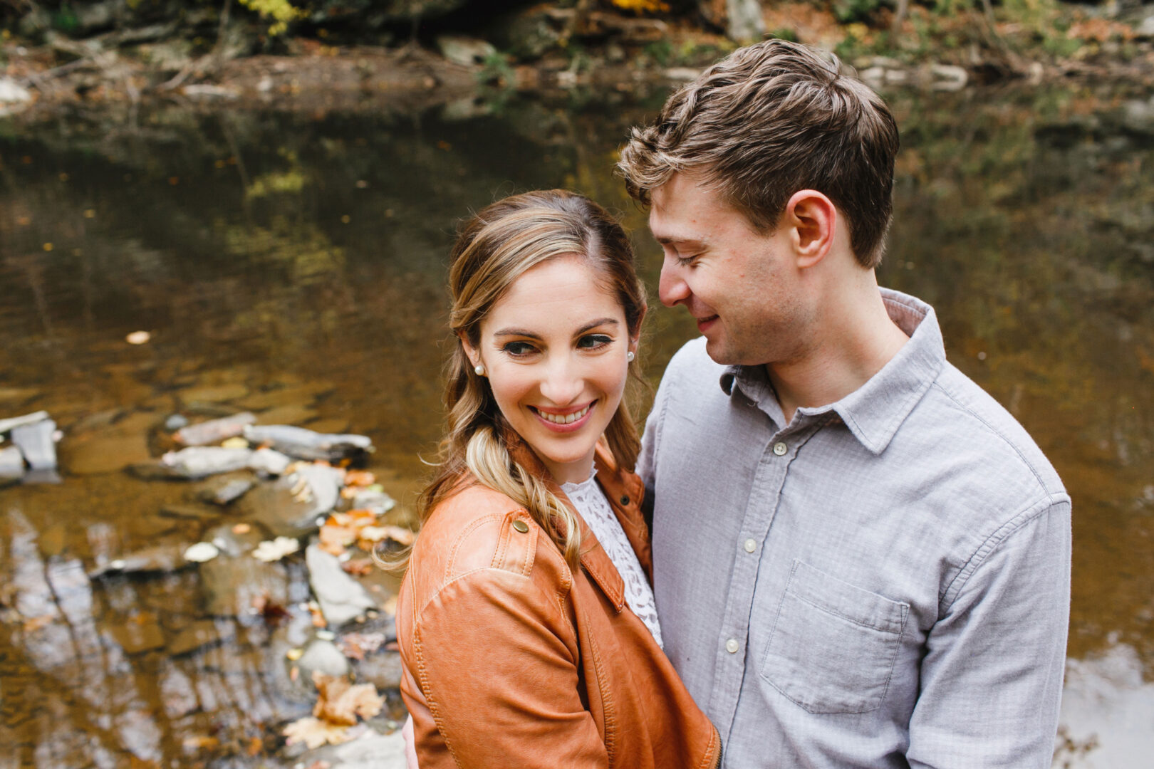 A couple stands close to each other by a calm river, surrounded by rocks and autumn leaves in Chestnut Hill. The woman is wearing a brown jacket, and the man is in a light gray shirt, sharing an intimate moment that captures their engagement's serene beauty.