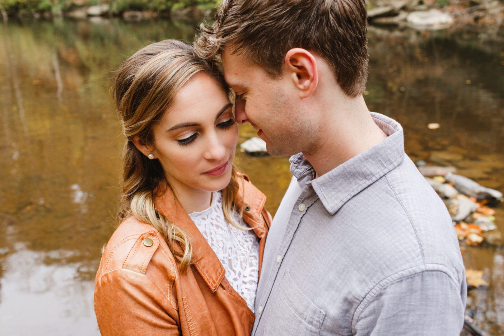 A couple stands closely by a calm body of water at Chestnut Hill, heads gently touching. The woman wears a brown jacket; the man wears a gray shirt, capturing their tender engagement moment.
