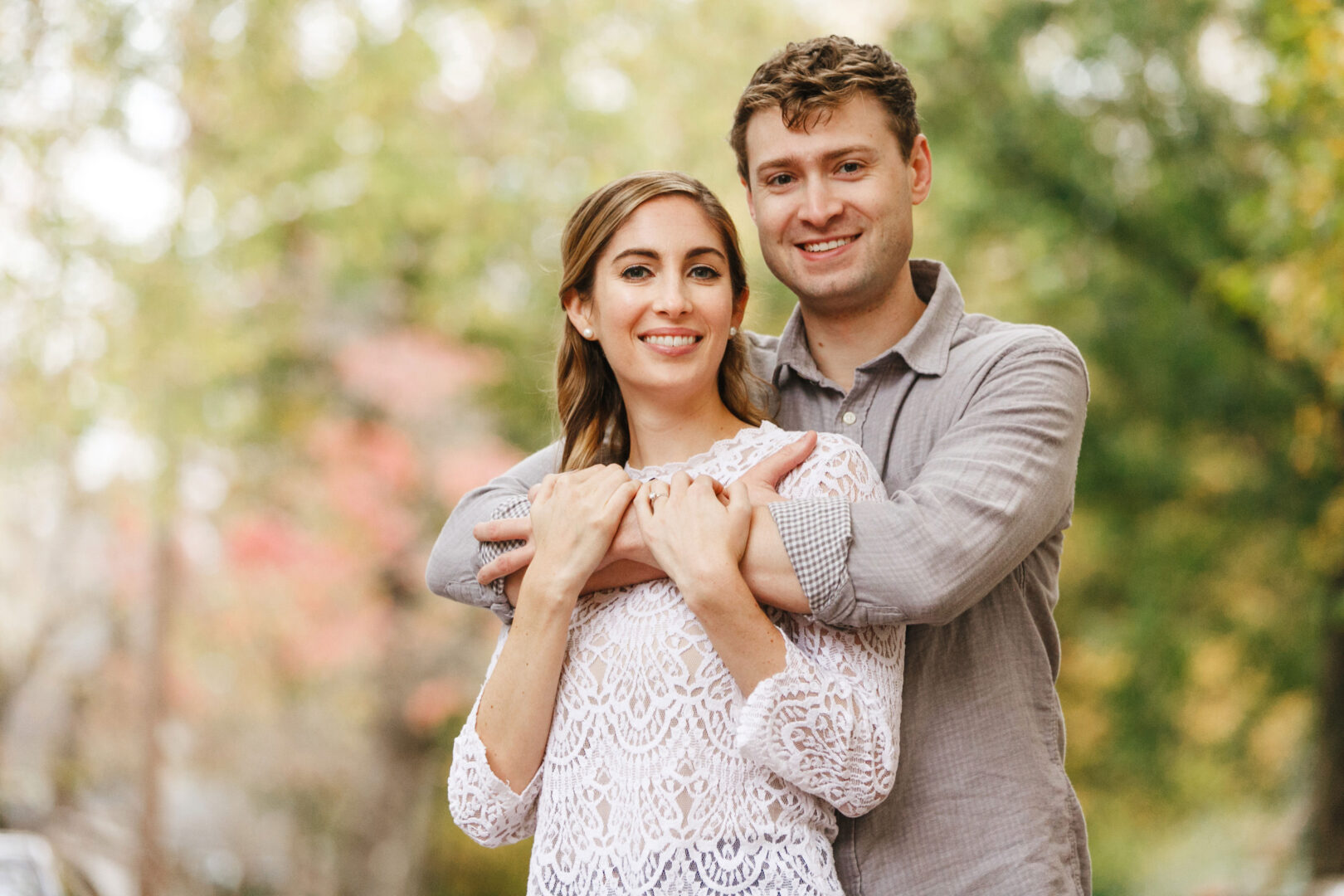 A man embraces a woman from behind, both smiling amidst the serene beauty of Chestnut Hill Engagement, with trees and blurred foliage creating a picturesque backdrop.
