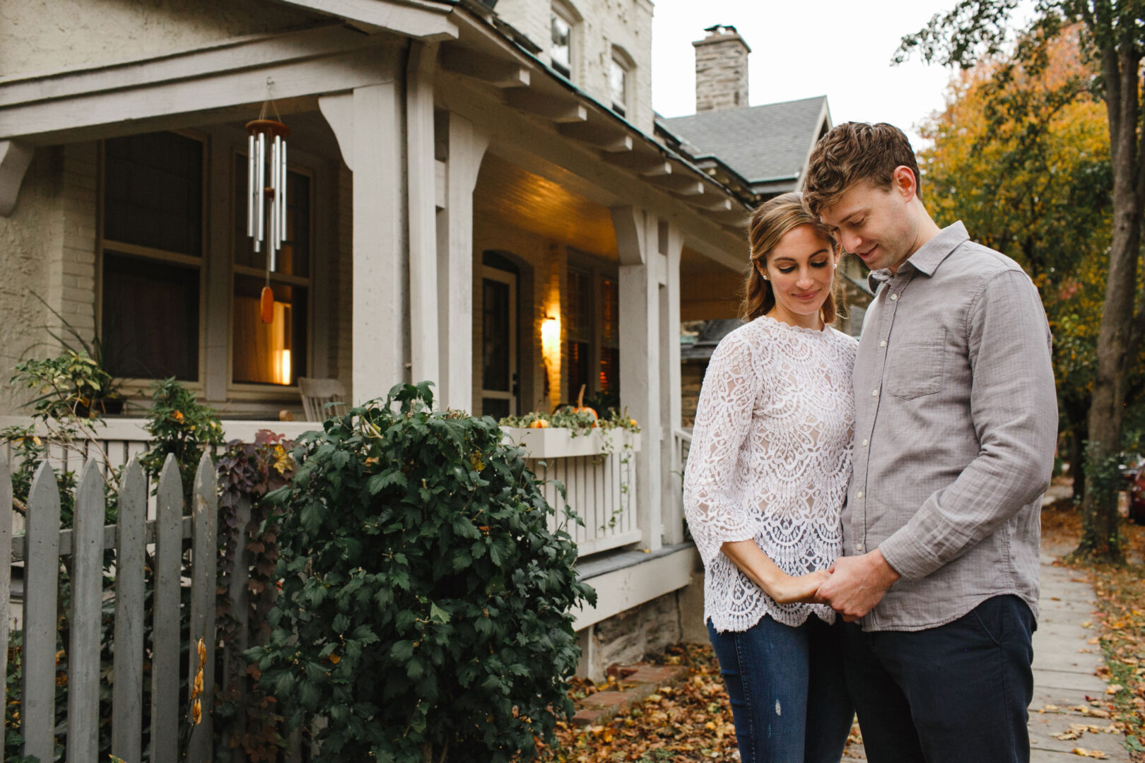 A couple stands together, holding hands and looking down, in front of a quaint Chestnut Hill house with a porch and vibrant autumn foliage, capturing their engagement beautifully.