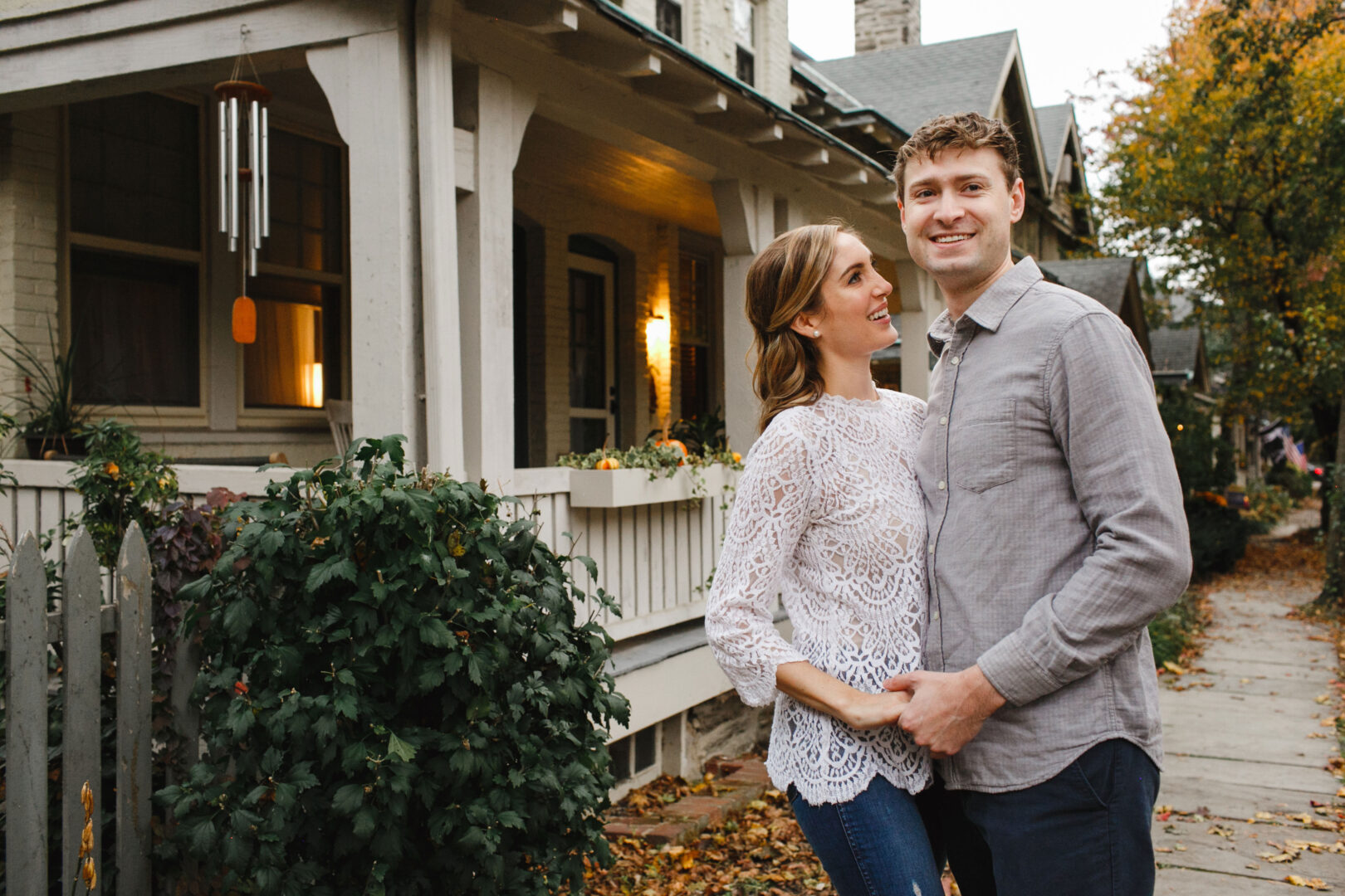 A couple stands together in front of a charming Chestnut Hill house with a porch, surrounded by greenery. The man wears a gray shirt; the woman is in a white lace top. Fall leaves scatter across the sidewalk, adding to their engagement's warm, picturesque setting.