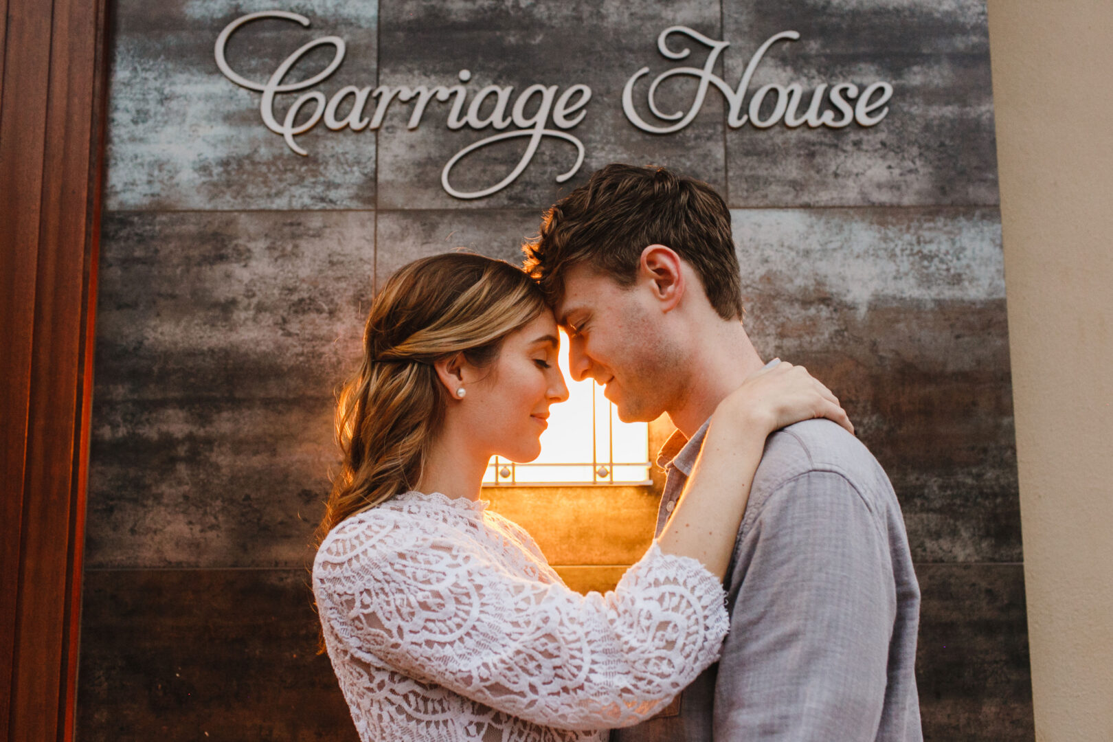 A couple embraces with foreheads touching in front of a wall that reads "Carriage House," capturing the essence of a Chestnut Hill engagement. A light fixture softly illuminates the tender moment in the background.
