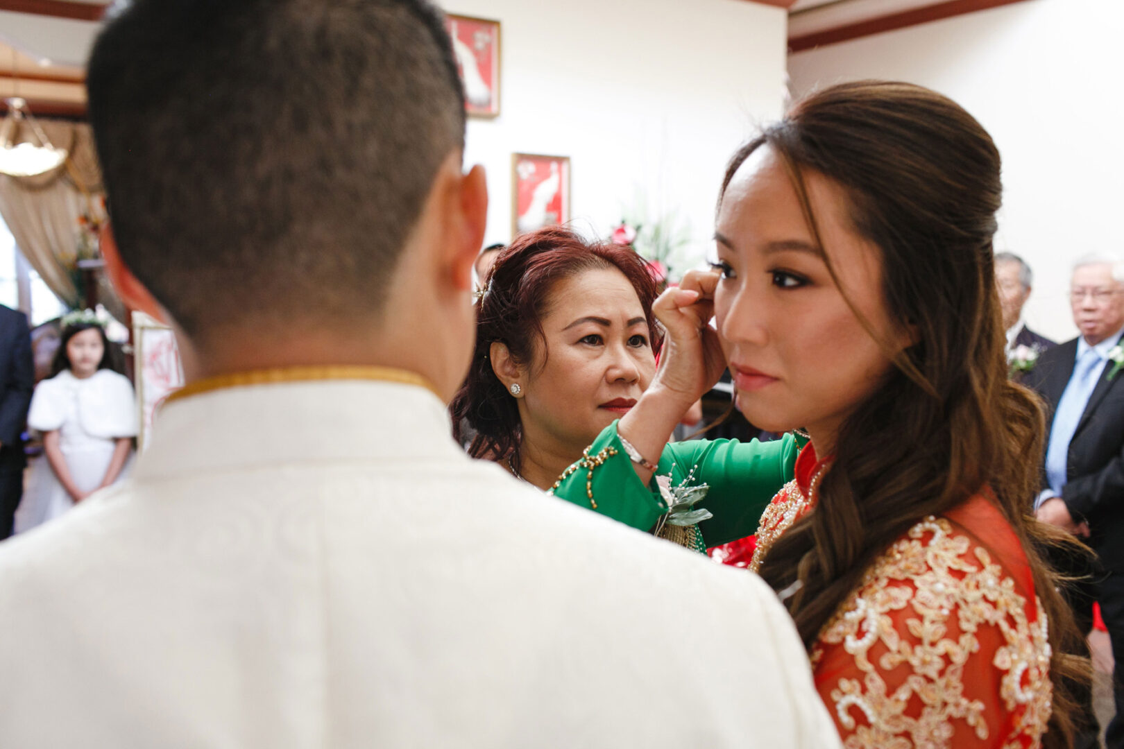 At the Hershey Wedding, a woman in a green dress adjusts the hair of the bride in a red gown, while the groom in a white suit stands nearby.