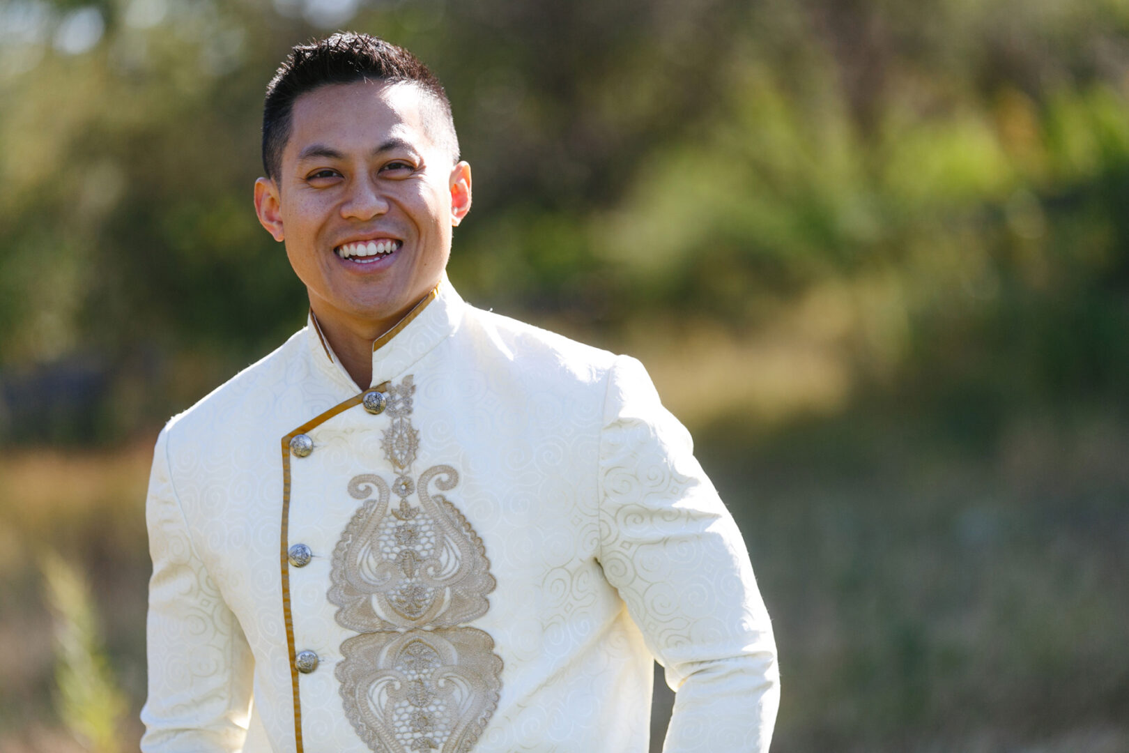 A person smiling broadly, wearing a light-colored suit with an intricate design on the front, stands outdoors at a Hershey wedding, surrounded by greenery in the blurred background.