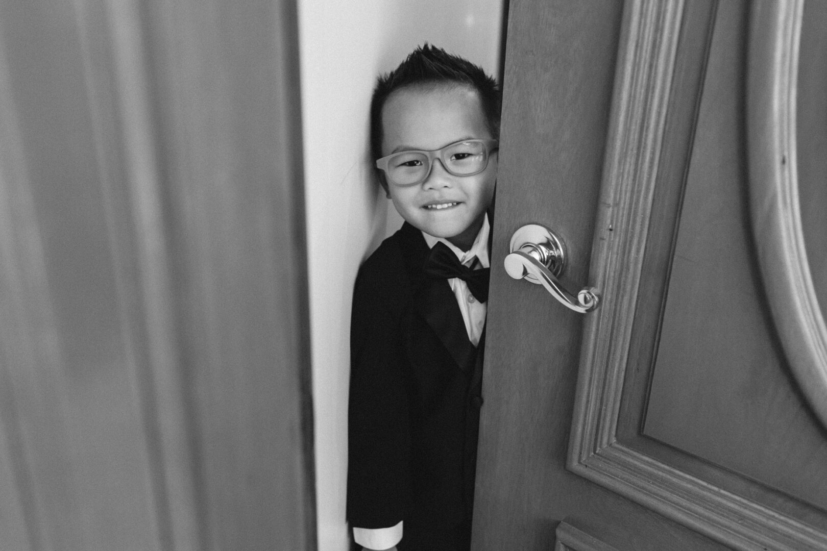 A young child in a suit and bowtie peeks through a slightly open wooden door, smiling with the excitement of a Hershey wedding in the air. The timeless image is captured beautifully in black and white.