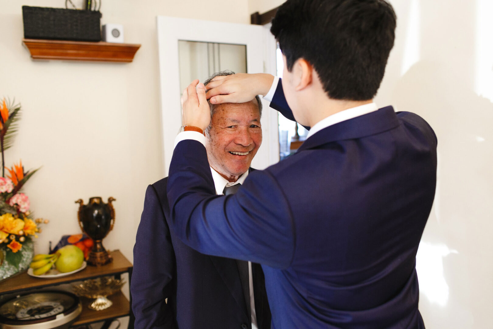 In a room adorned with decorative items and fruit on the table, a man in a suit gently adjusts an older gentleman's hair, preparing for the elegant Hershey wedding.