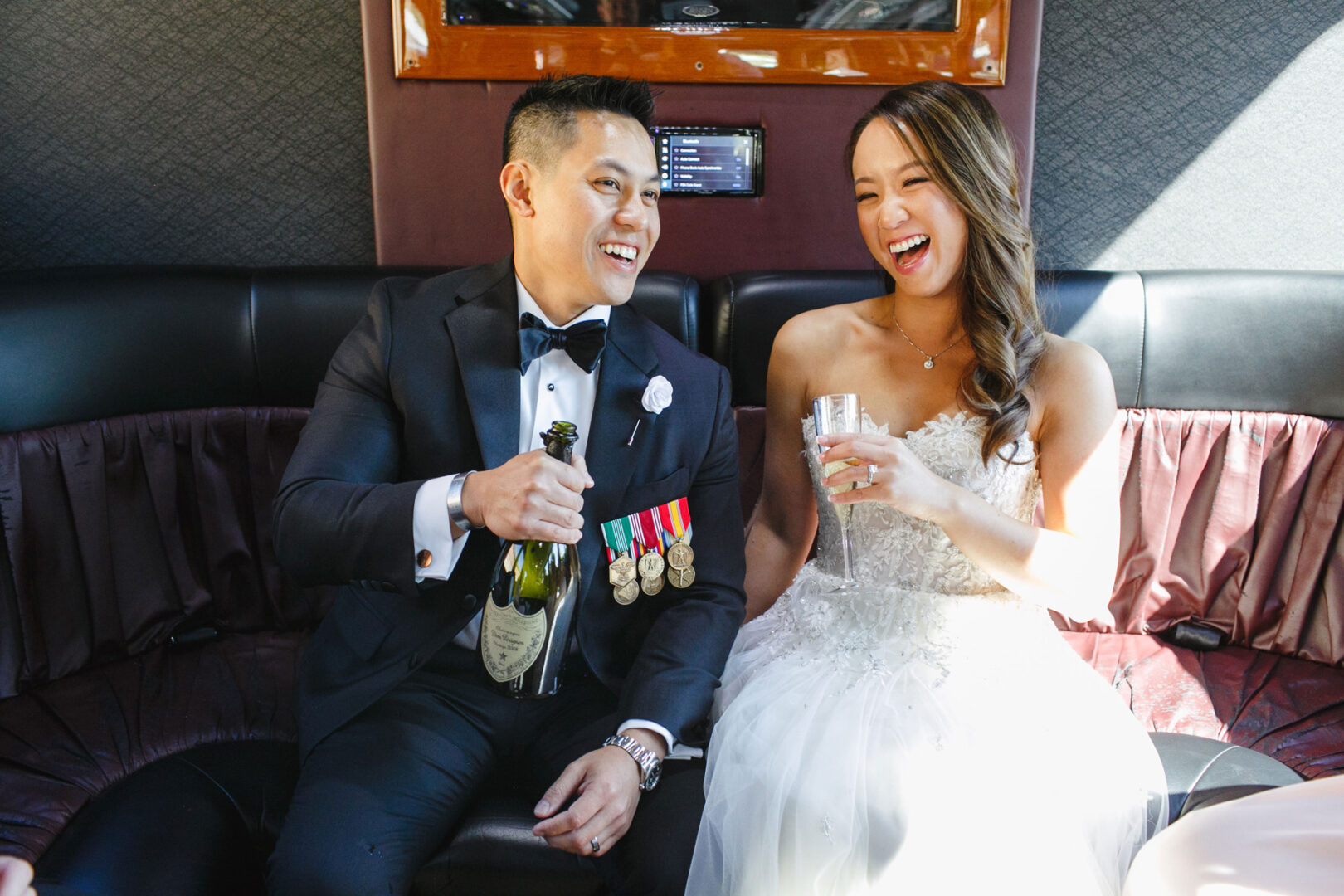 In a well-lit room, the bride and groom, dressed in elegant formal attire, sit on a couch with joyful smiles, holding champagne bottles. It's the perfect Hershey wedding moment, capturing their happiness and love.