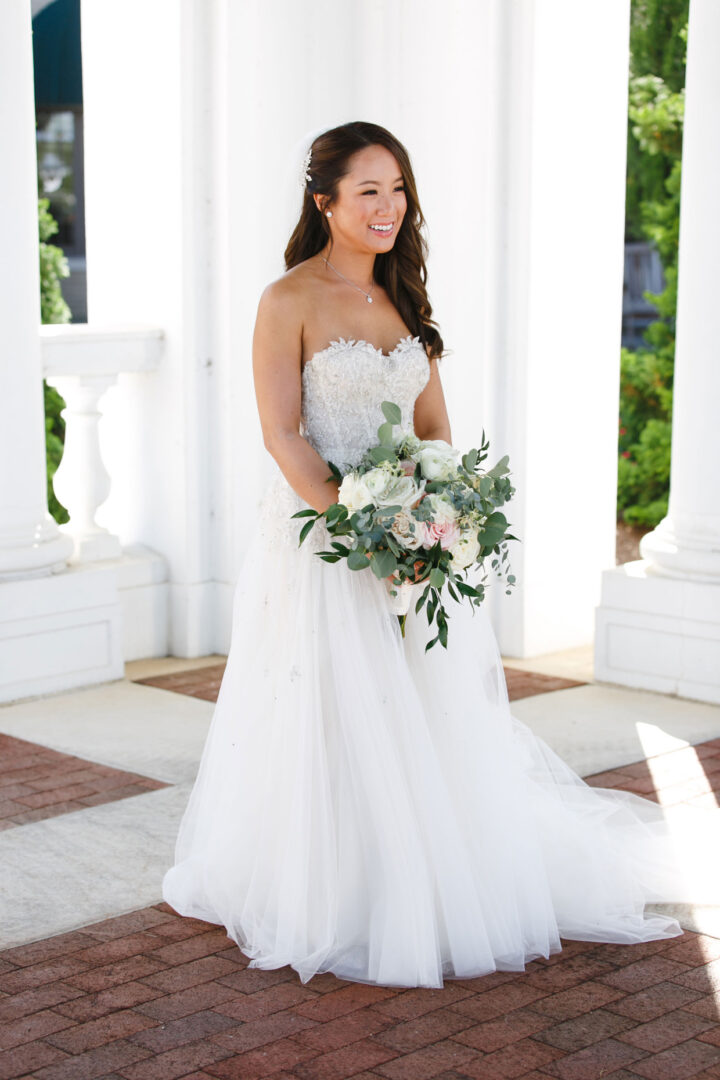 Bride in a strapless white wedding gown holding a bouquet of white and pink flowers, standing gracefully in front of classic white columns and lush greenery at the enchanting Hershey Wedding venue.