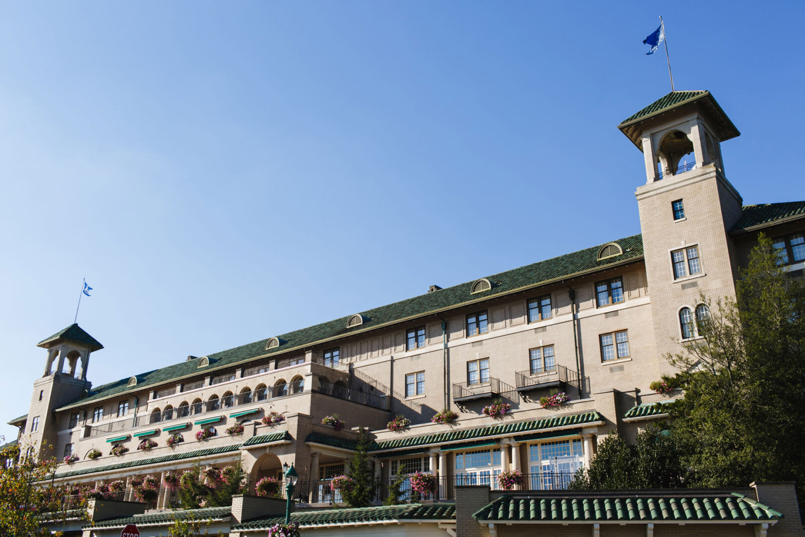 The exterior of a large historic building, perfect for a Hershey wedding, features green roofs and arched windows. A flag graces the top while balconies adorned with potted plants add charm. All this under a clear blue sky creates a picturesque setting for your special day.