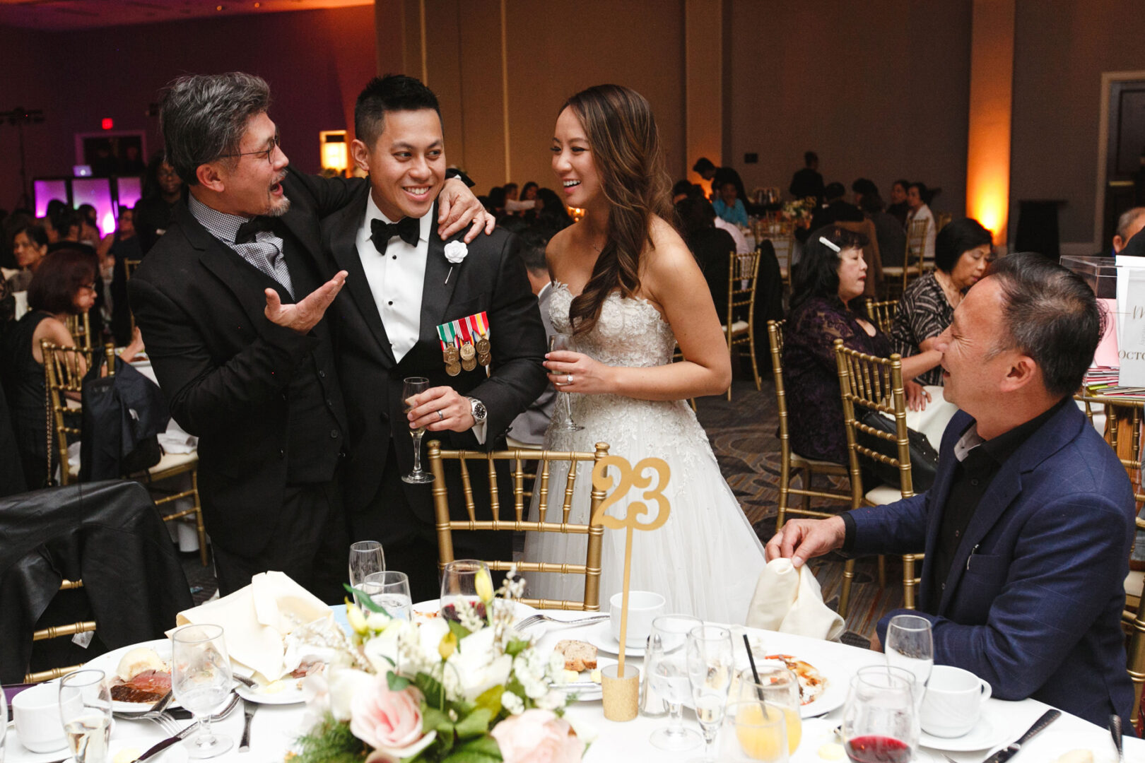 A smiling couple in formal attire stand beside a seated man at the elegant Hershey wedding. Another man, also standing, gestures animatedly near the couple. The table is adorned with floral arrangements and drinks, adding to the celebratory atmosphere.