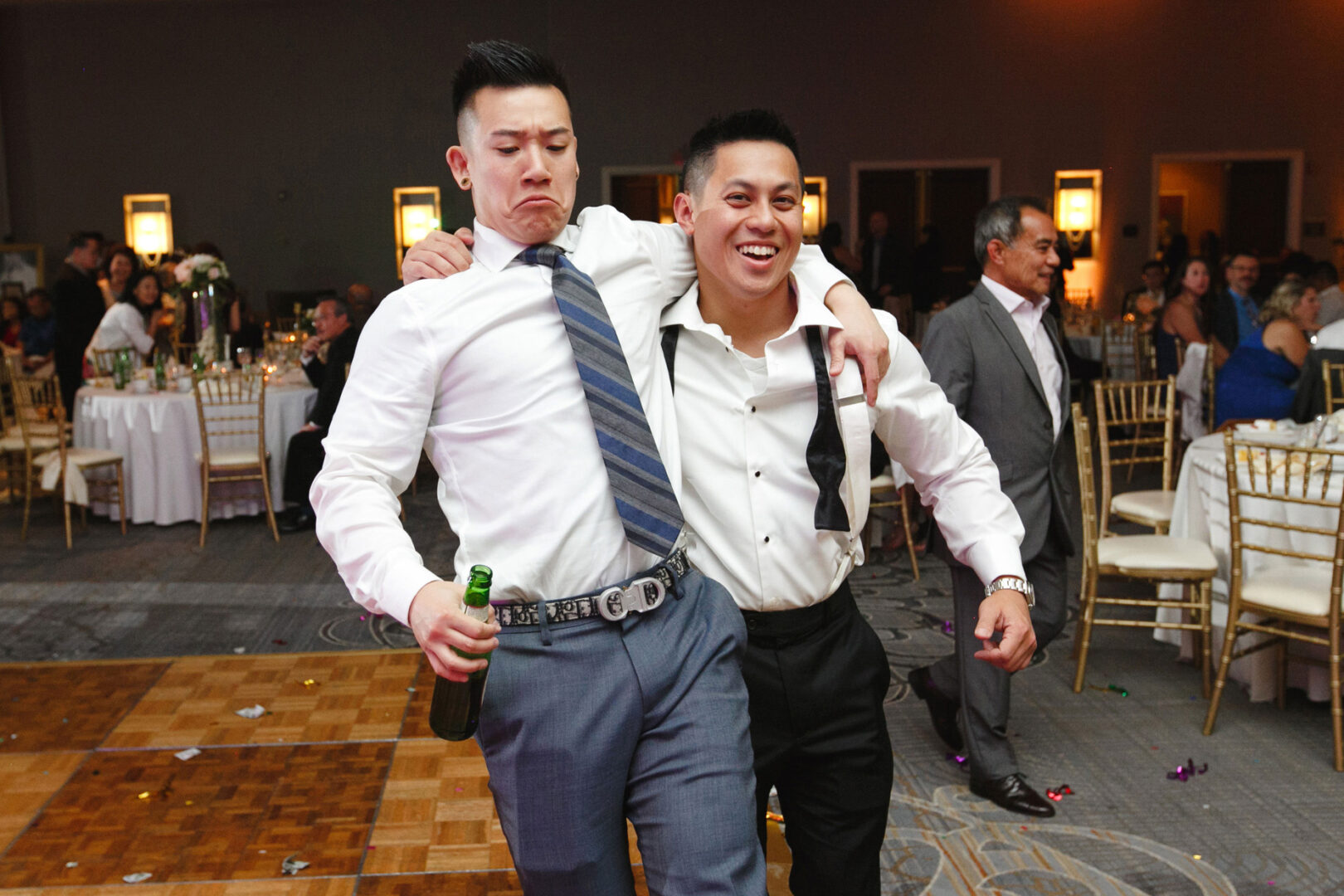Two men, smiling and walking arm-in-arm at a Hershey Wedding event. One holds a beer bottle while decorated tables and seated guests create a festive background.