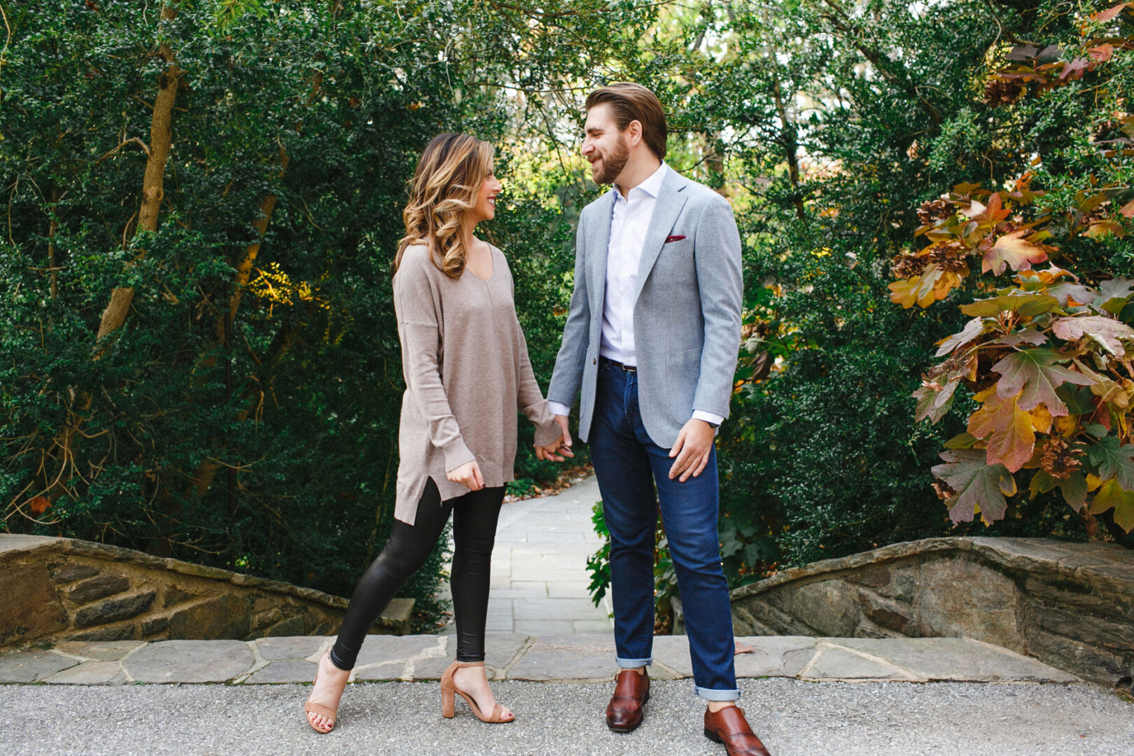 A couple stands on an outdoor path at Longwood Gardens, surrounded by greenery, holding hands and smiling at each other. The woman wears a beige sweater and black pants, while the man sports a light blazer and jeans—a perfect setting for an engagement session.