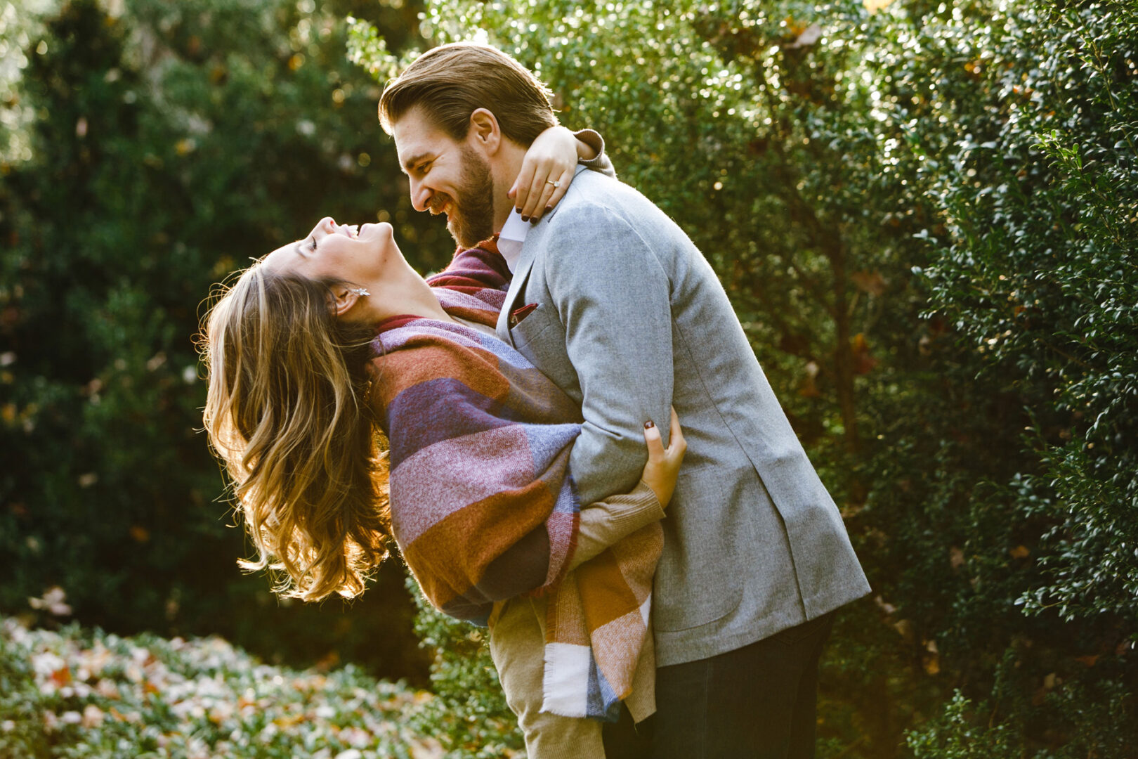 A couple embraces and smiles at each other in the garden, capturing the magic of their Longwood Gardens engagement session. The woman is wrapped in a colorful shawl, while the man sports a sleek gray jacket, adding to the serene beauty of their cherished moment.