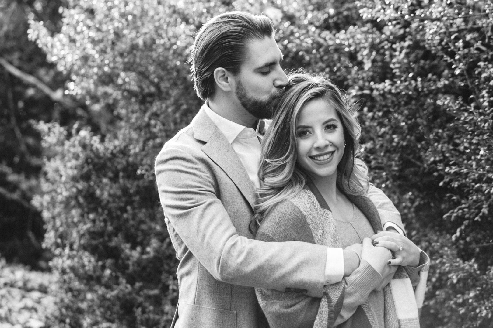 In a timeless black and white image, captured during a Longwood Gardens engagement session, a man tenderly kisses the head of a smiling woman while hugging her from behind. Both wear light-colored clothing, their happiness beautifully immortalized in the serene garden setting.