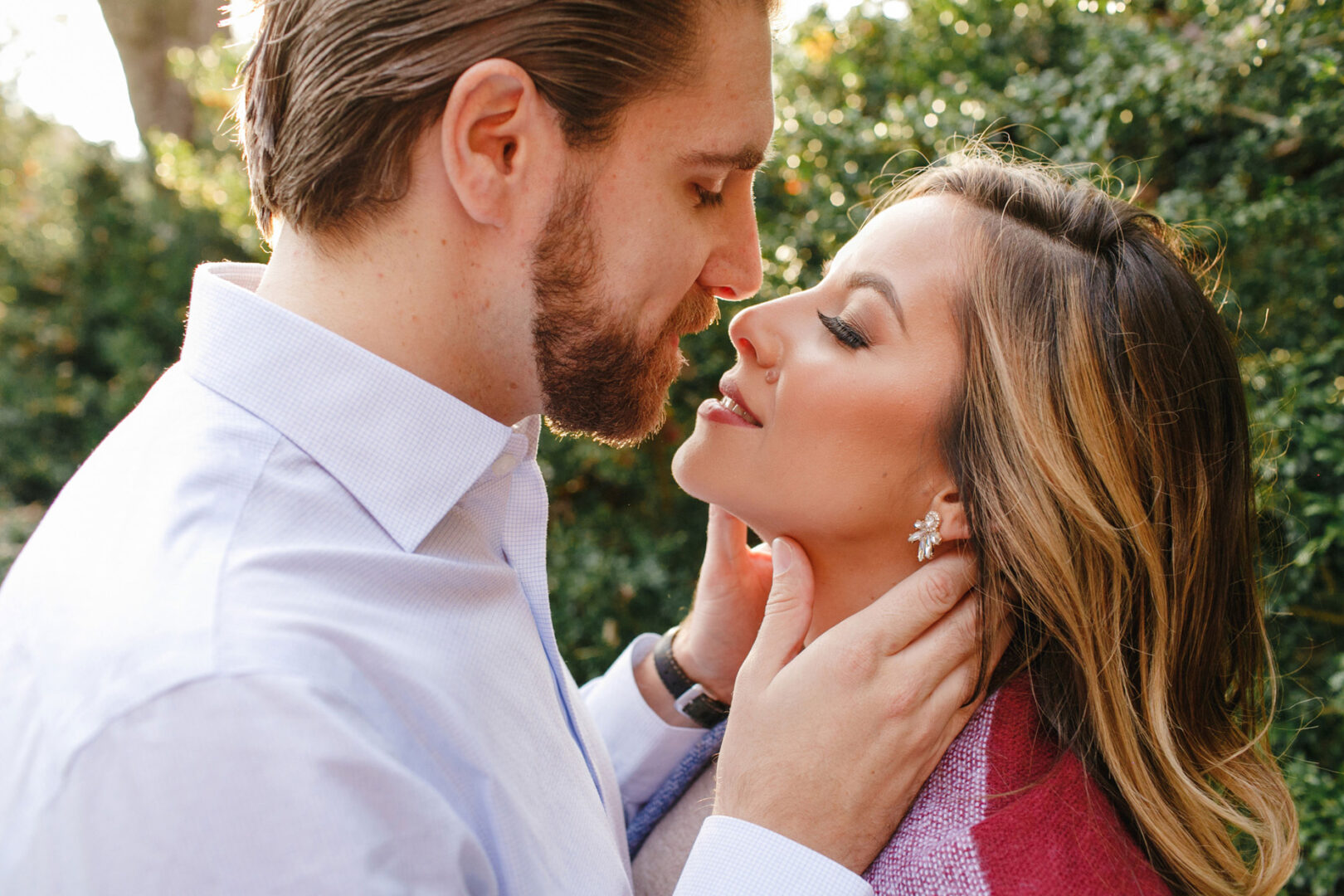 Amidst the lush greenery of Longwood Gardens, a couple shares an intimate moment during their engagement session. The man tenderly holds the woman's face with both hands as they gaze at each other closely, enveloped by nature's serene beauty.