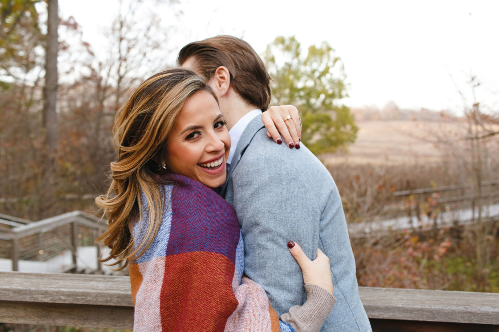 During a Longwood Gardens engagement session, a woman smiles warmly as she hugs her partner on a wooden walkway, surrounded by nature's serene beauty.