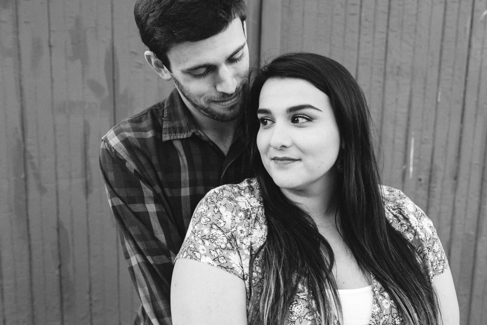 In a relaxed setting against a wooden background, a man and woman stand closely together, embodying a serene Valley Forge engagement. The man is slightly behind the woman as they share a quiet moment filled with intimate connection.