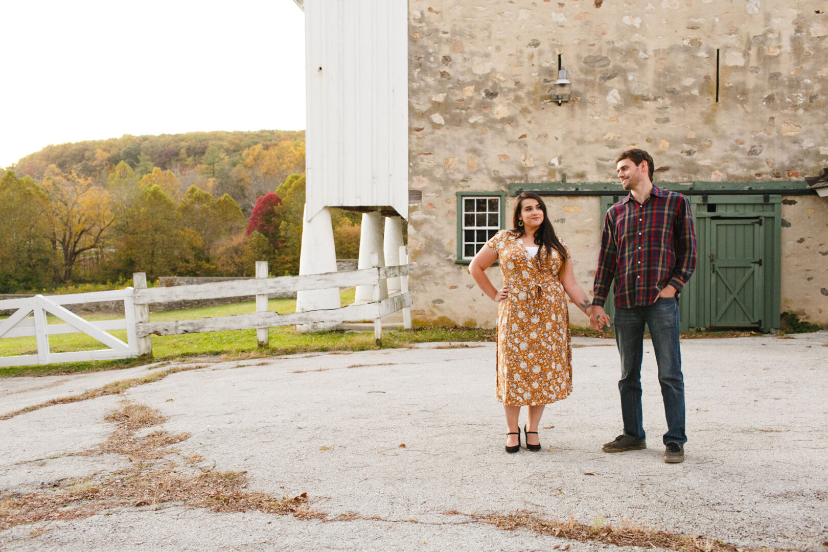 At Valley Forge, a couple holding hands stands on a concrete area next to a rustic stone building. The woman wears a yellow floral dress, and the man wears a plaid shirt and jeans. Tall trees frame this engagement scene in the background.