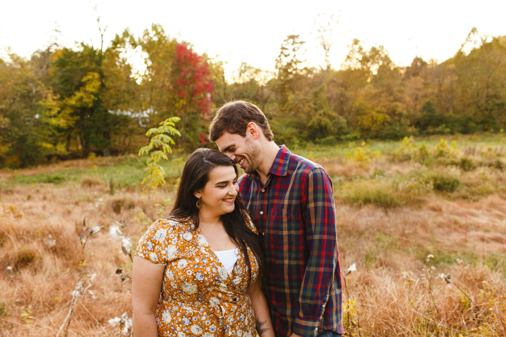 A couple stands in a field at Valley Forge, surrounded by vibrant autumn foliage. The woman in a floral dress and the man in his plaid shirt share a loving gaze, capturing the essence of their engagement.