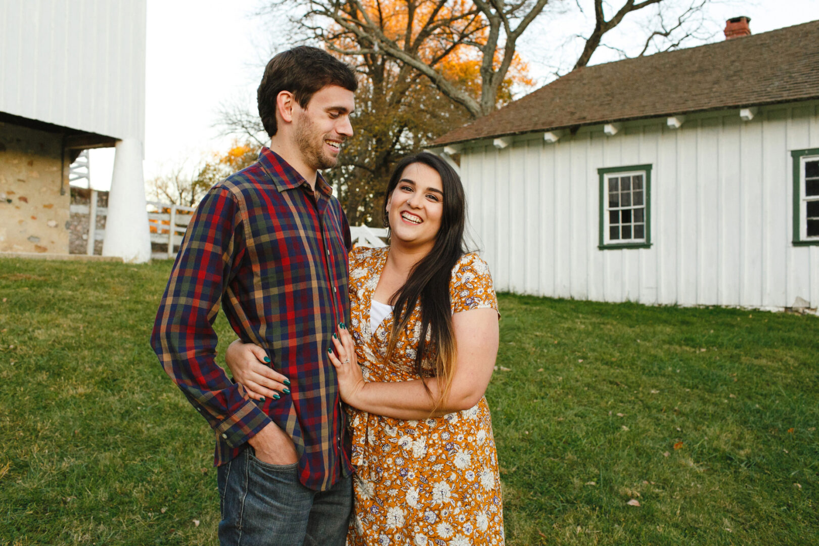 A man and woman stand smiling on a grassy lawn at Valley Forge, with white buildings and trees in the background, capturing their joyful engagement moment.