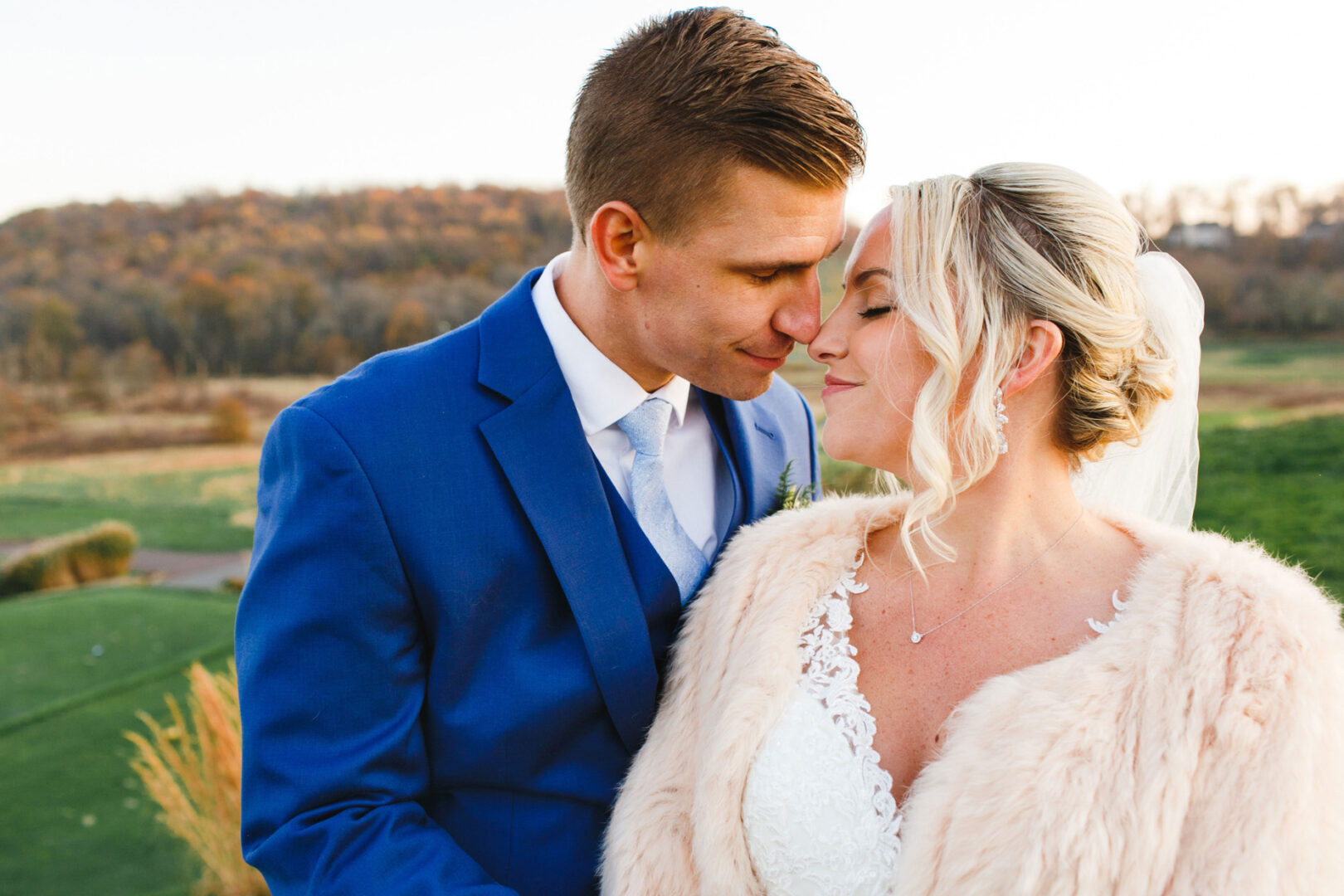 A couple in formal attire, with the man in a blue suit and the woman in a white dress and fur shawl, stand close together outdoors at their French Creek Golf Club wedding. A scenic, blurred landscape is in the background.