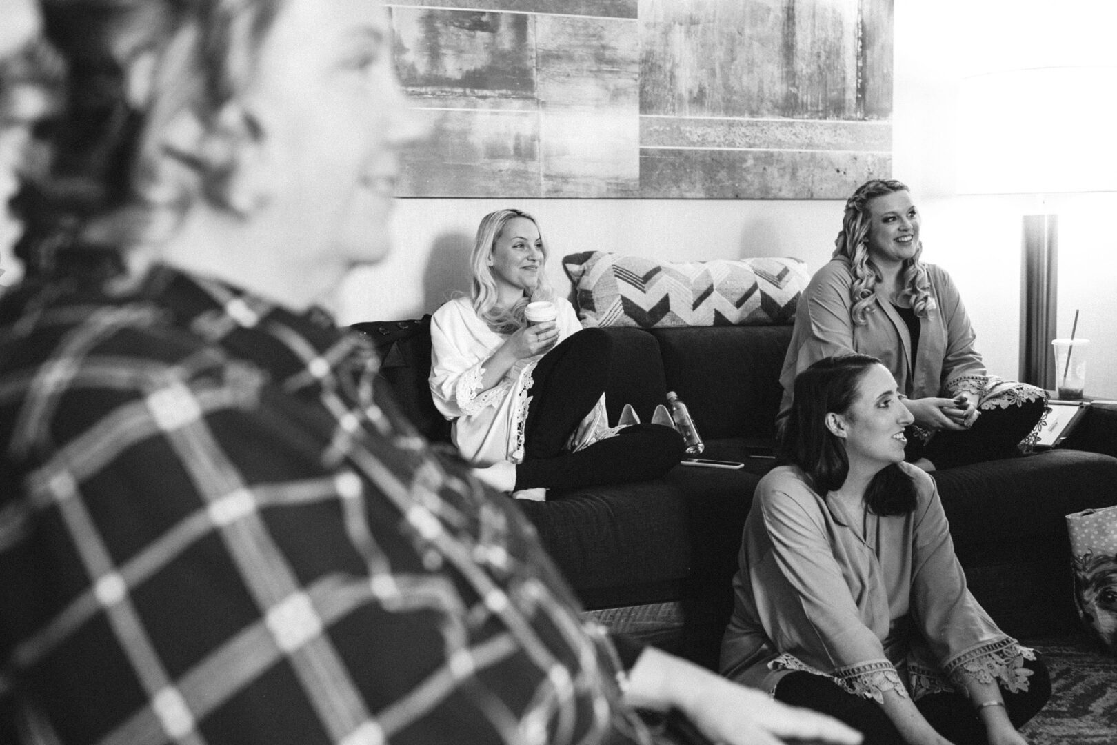 Four women gather in a cozy living room, three perched on a couch while one relaxes on the floor. The space is accented by a large abstract painting and an elegant floor lamp, creating an ambiance reminiscent of the refined charm found at a French Creek Golf Club Wedding.