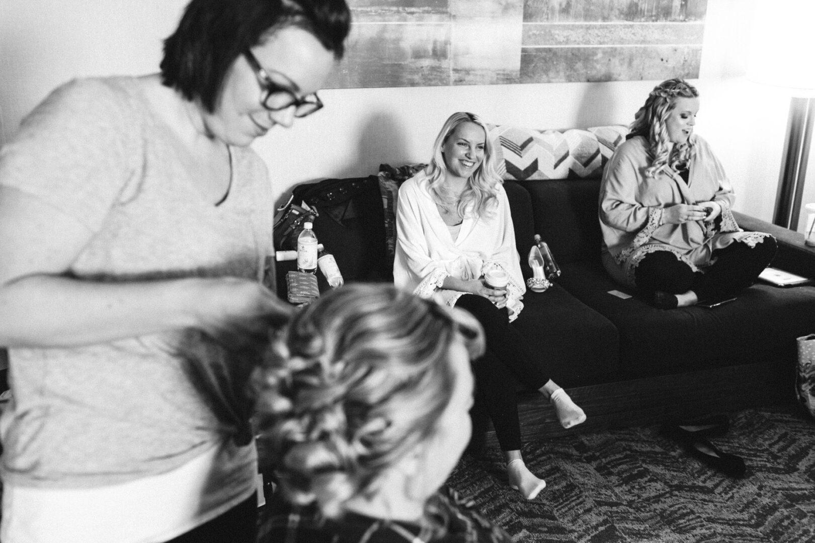 In a cozy room, where a woman styles another's hair, three seated women relax on a couch, reminiscing about the unforgettable French Creek Golf Club wedding they attended.