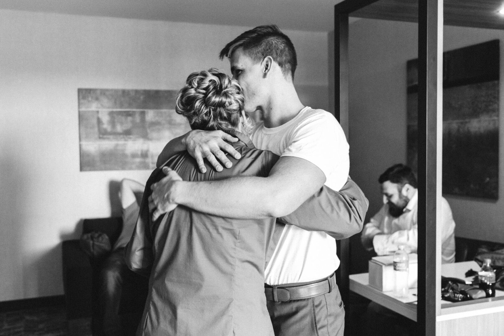 In a cozy room, a man in a white shirt lovingly embraces a woman in a jacket, capturing the warmth of the moment, reminiscent of joyous celebrations like those at French Creek Golf Club Wedding. Meanwhile, another man sits quietly at a desk in the background.