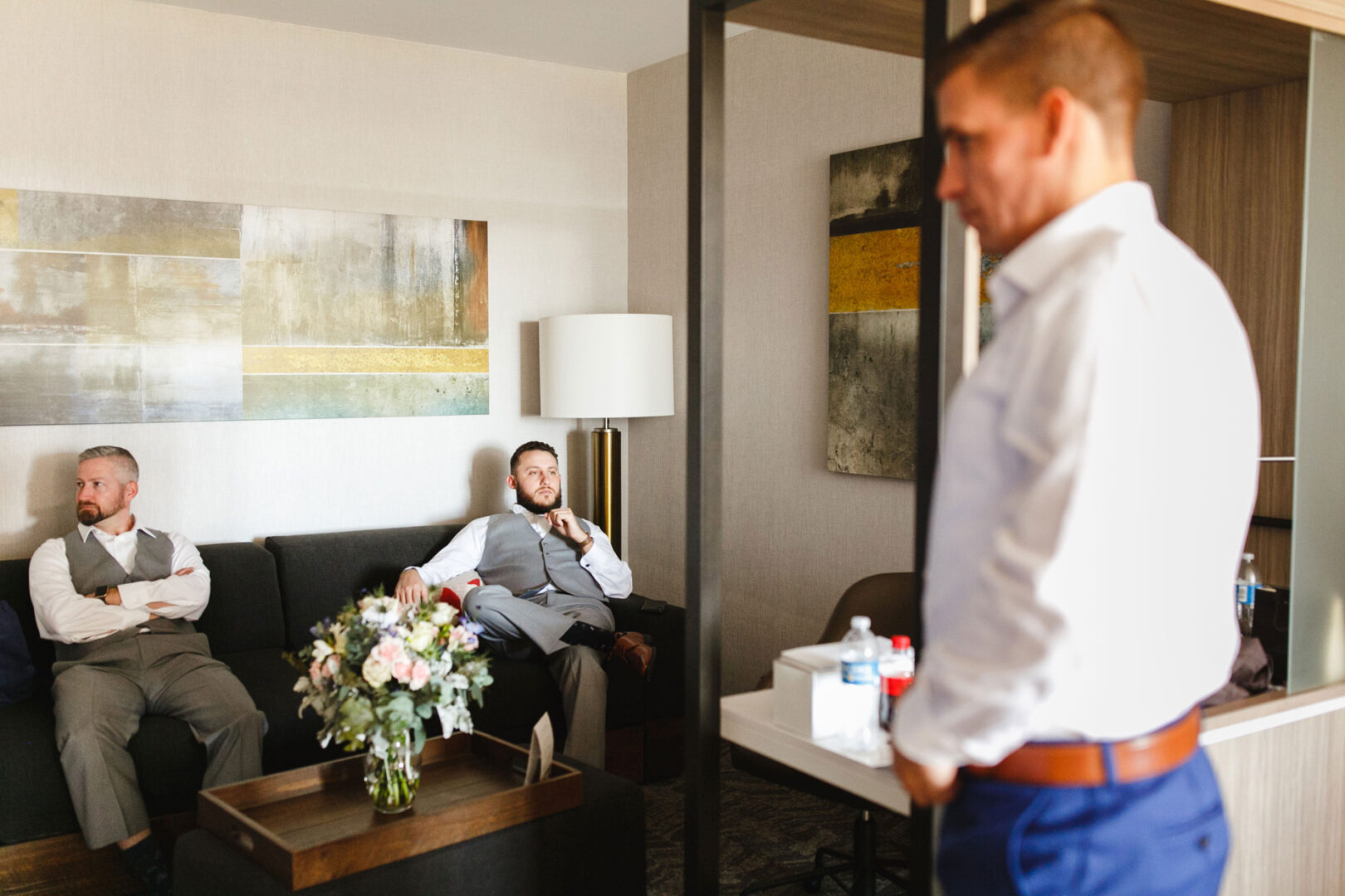 Three men in a room, two sitting on a couch in suits and one standing in the foreground, exude an air of elegance reminiscent of a French Creek Golf Club wedding. A bouquet of flowers graces the table, adding a touch of refined charm.
