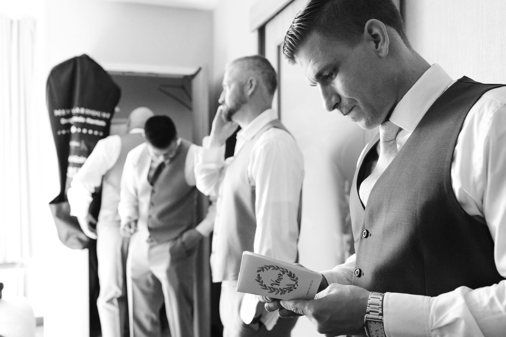 A group of men in suits, one reading a note, another on the phone discussing details for a French Creek Golf Club wedding, stand in a room with a garment bag hanging in the background.