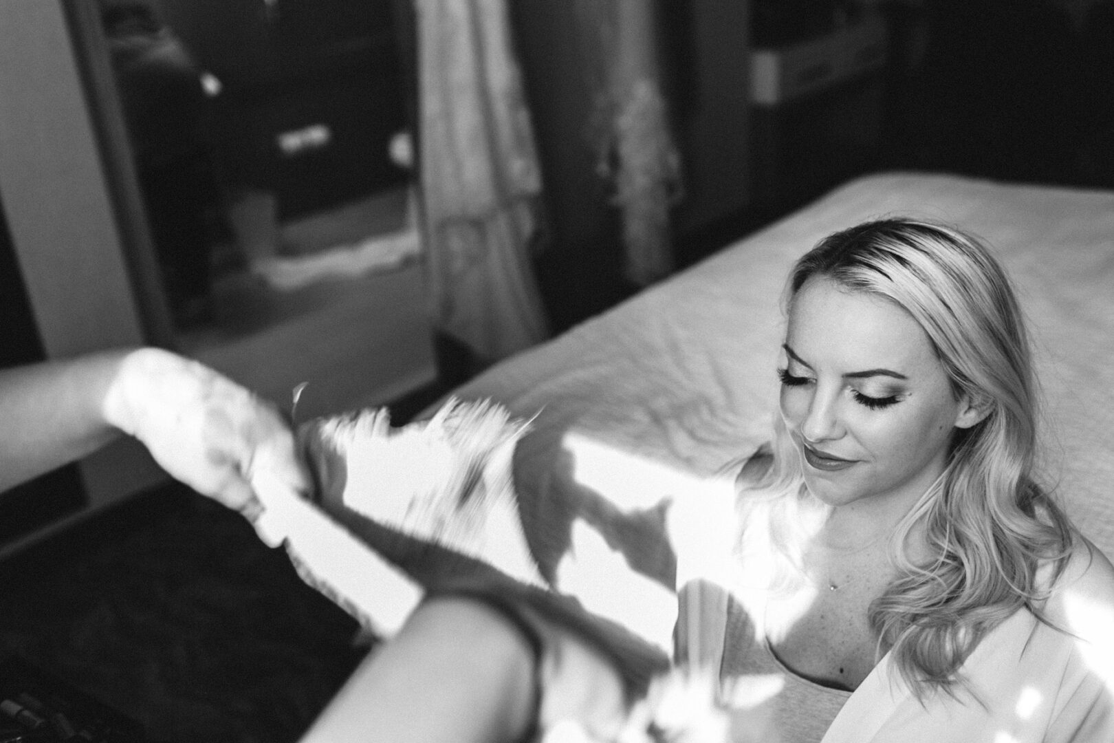 In a black and white photo, a blonde-haired woman sits gracefully on her bed, lost in thought. In the foreground, an unseen hand tenderly holds a piece of fabric. The scene evokes the quiet elegance of a French Creek Golf Club wedding morning.