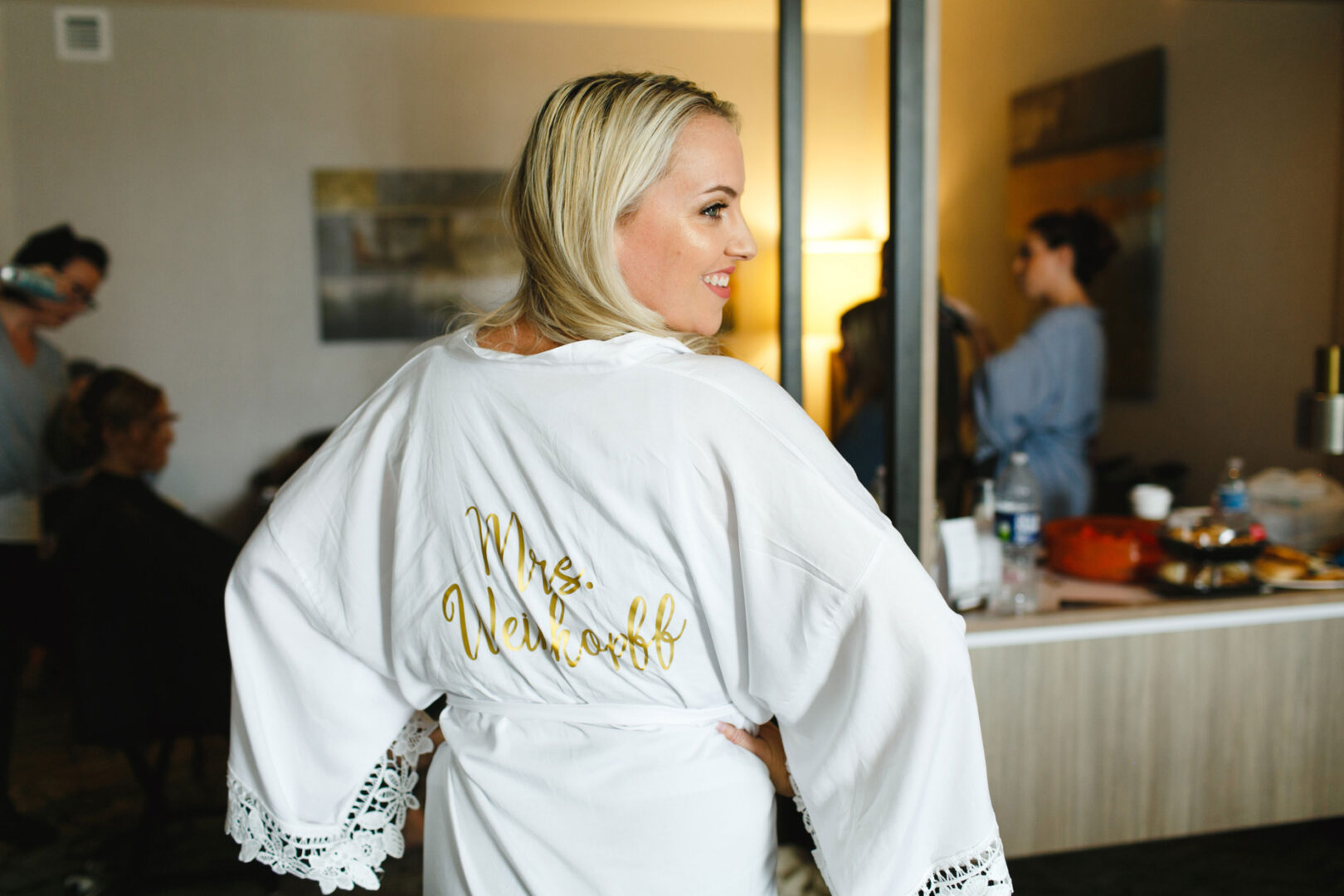 Woman in a white robe with "Mrs. Newhoff" on the back smiles, standing in a room buzzing with excitement at French Creek Golf Club, where wedding guests mingle in the background.